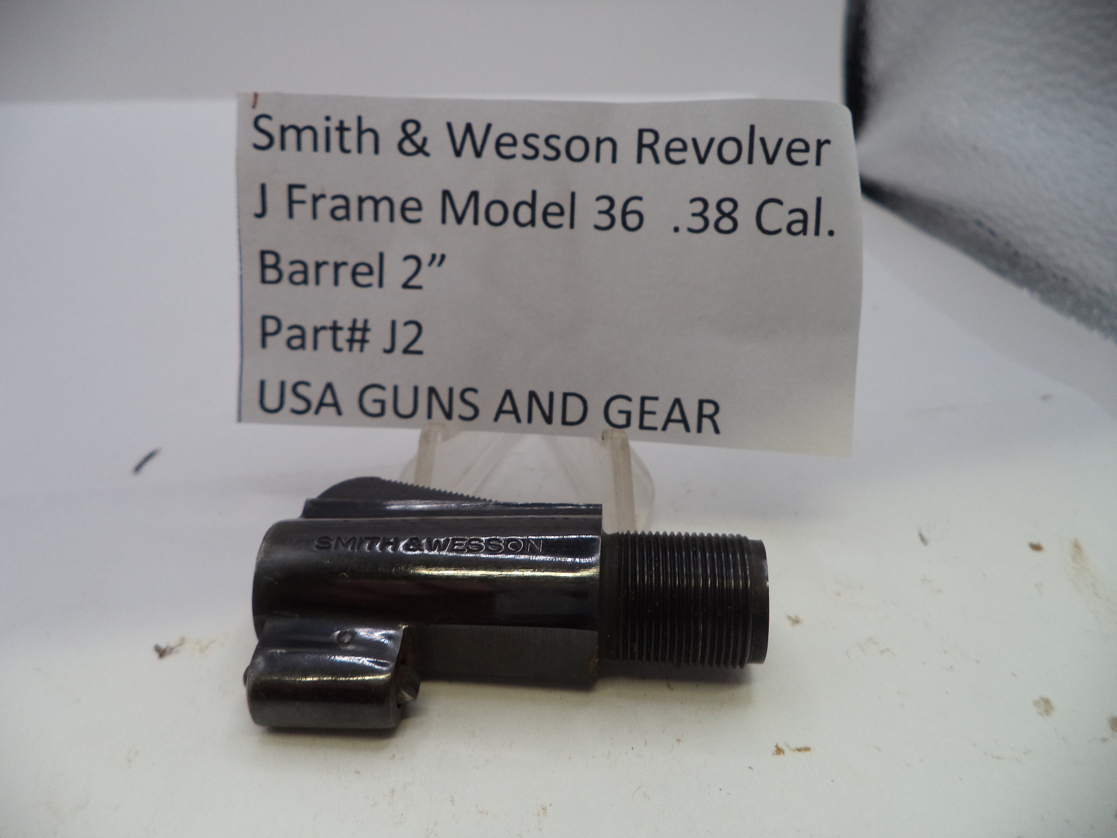 A black metal Smith & Wesson revolver barrel replacement part with a threaded section, displayed on a white surface with a handwritten paper label in the background reading 'Smith & Wesson Revolver J Frame Model 36 .38 Cal.