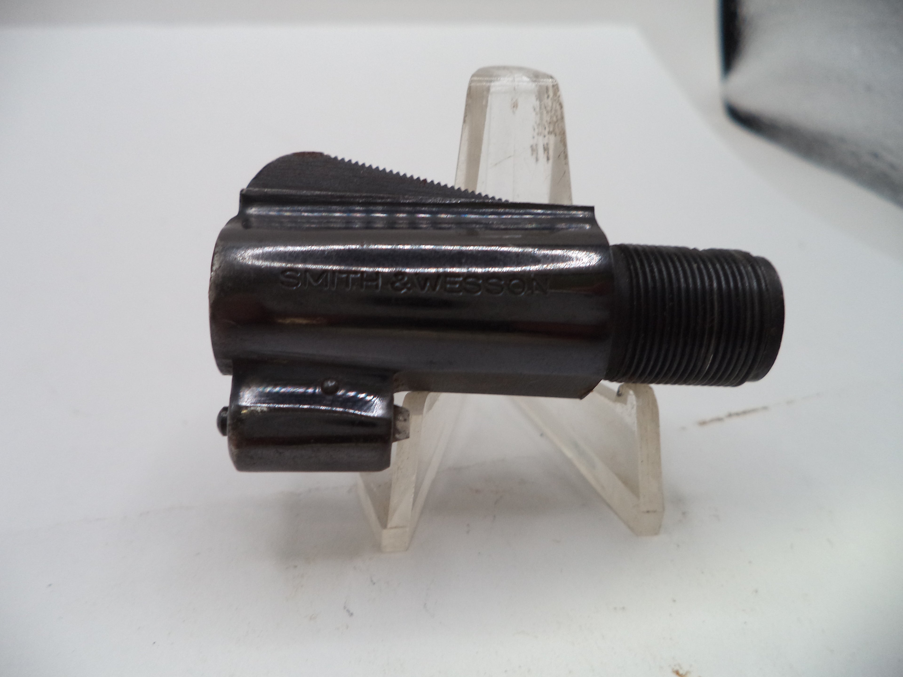 A black metal Smith & Wesson revolver cylinder block or barrel component with a threaded end, displayed on a clear plastic stand against a white background.