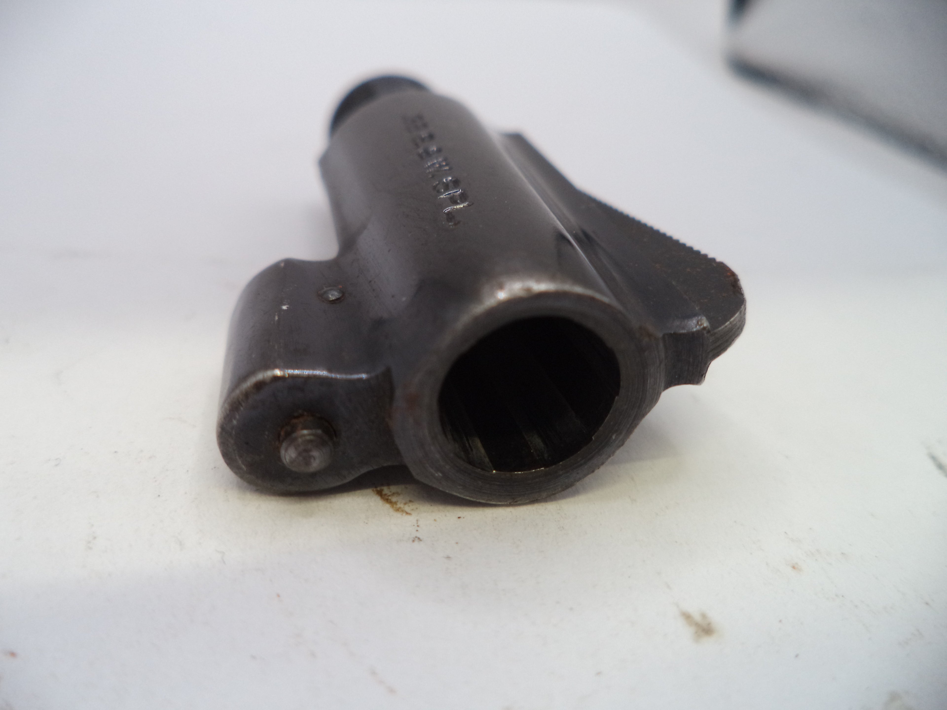 A close-up shot of a dark metal firearm component, specifically a receiver or barrel assembly with a visible bolt hole, resting on a white surface.