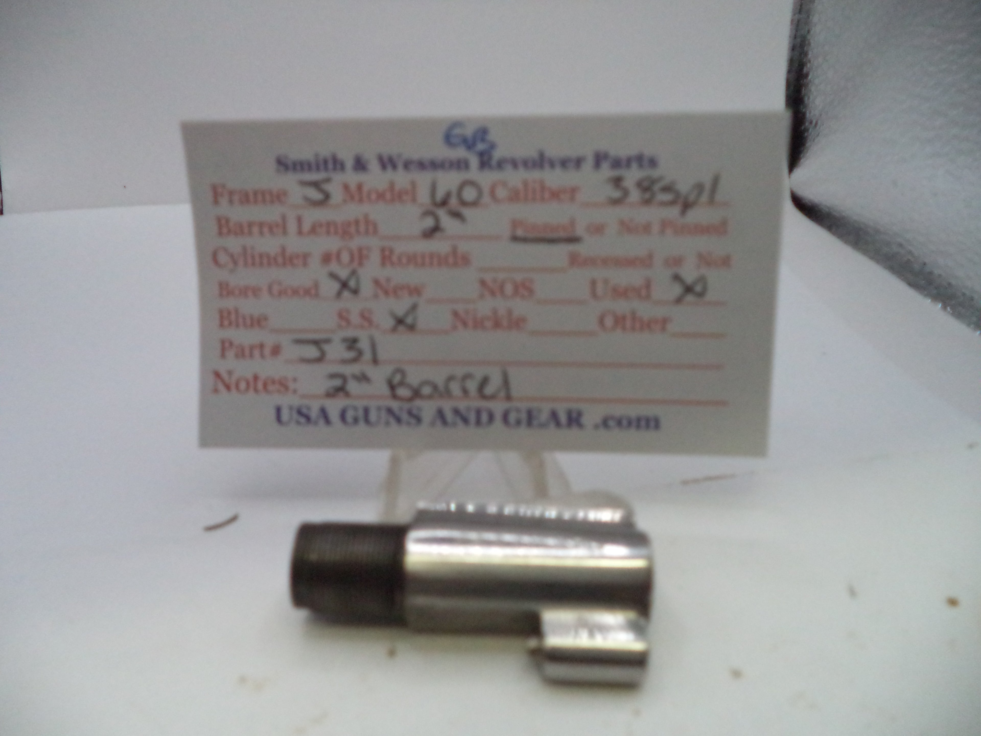 A used metal revolver part, likely a cylinder or frame component, displayed in front of a handwritten paper label card for Smith & Wesson revolver parts.