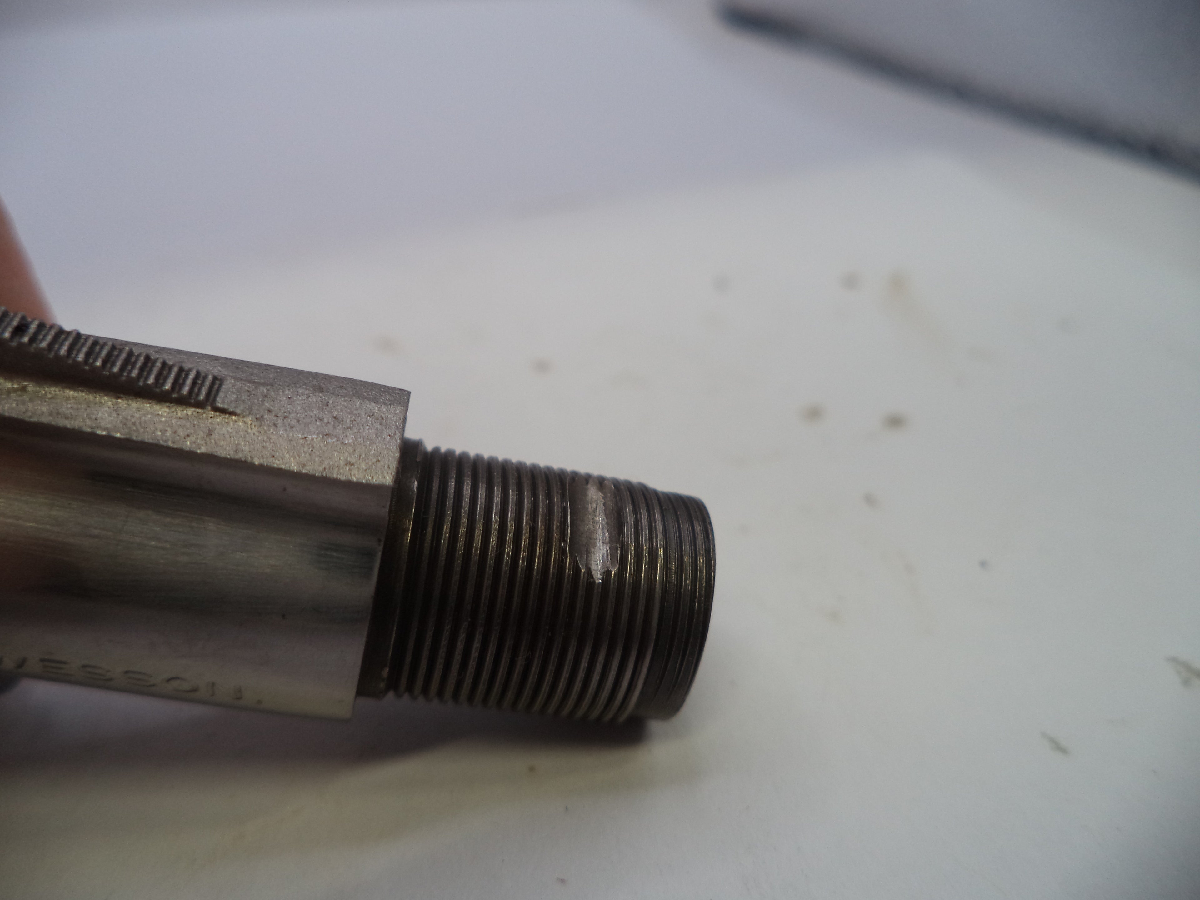 A close-up of a used metal threaded component, likely a mechanical fitting or adapter, held by a finger on a white surface.