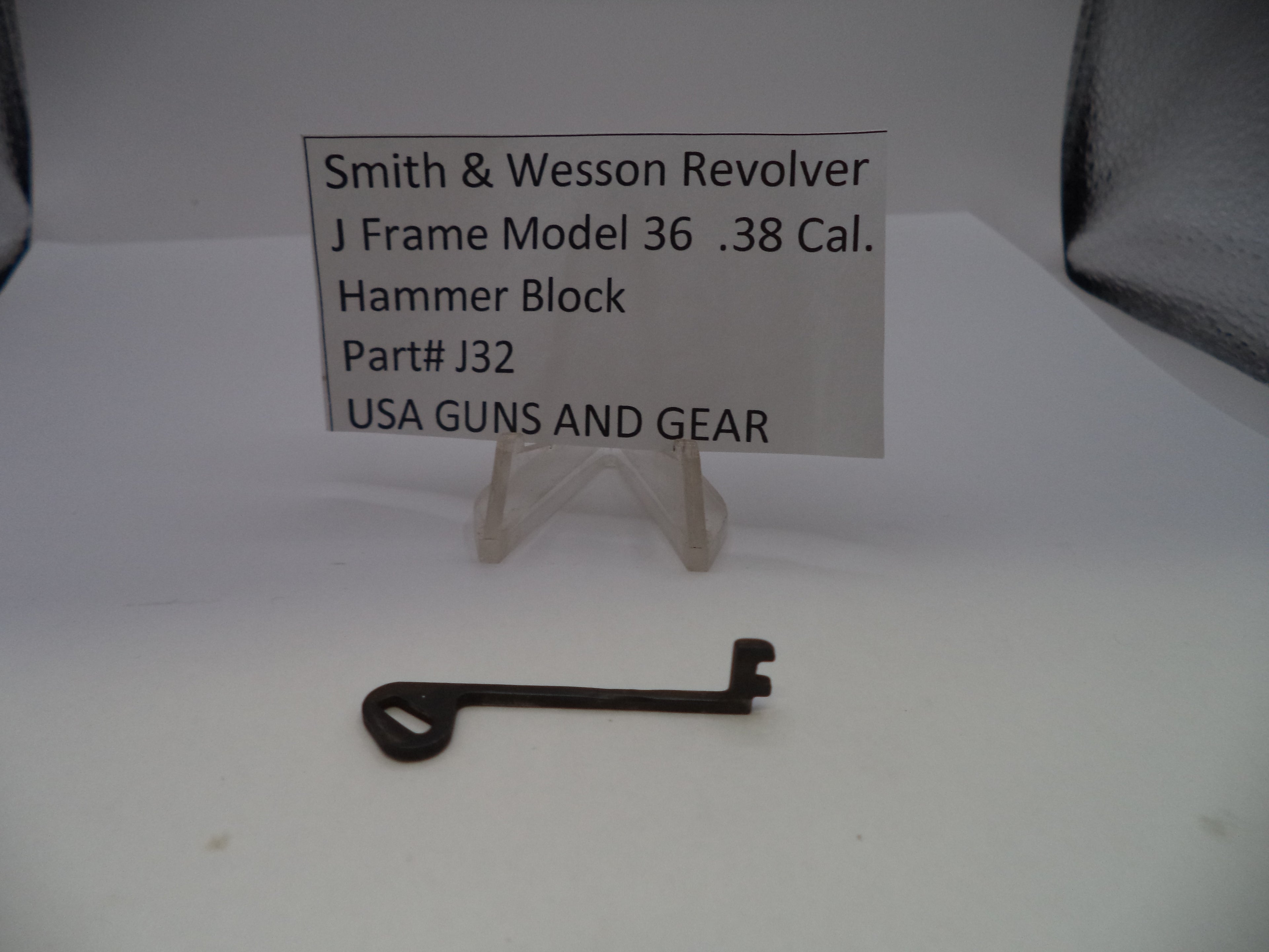 A small metal hammer block key for a Smith & Wesson revolver, displayed on a white surface with a clear plastic stand holding a label card that reads 'Smith & Wesson Revolver J Frame Model 36 .38 Cal.