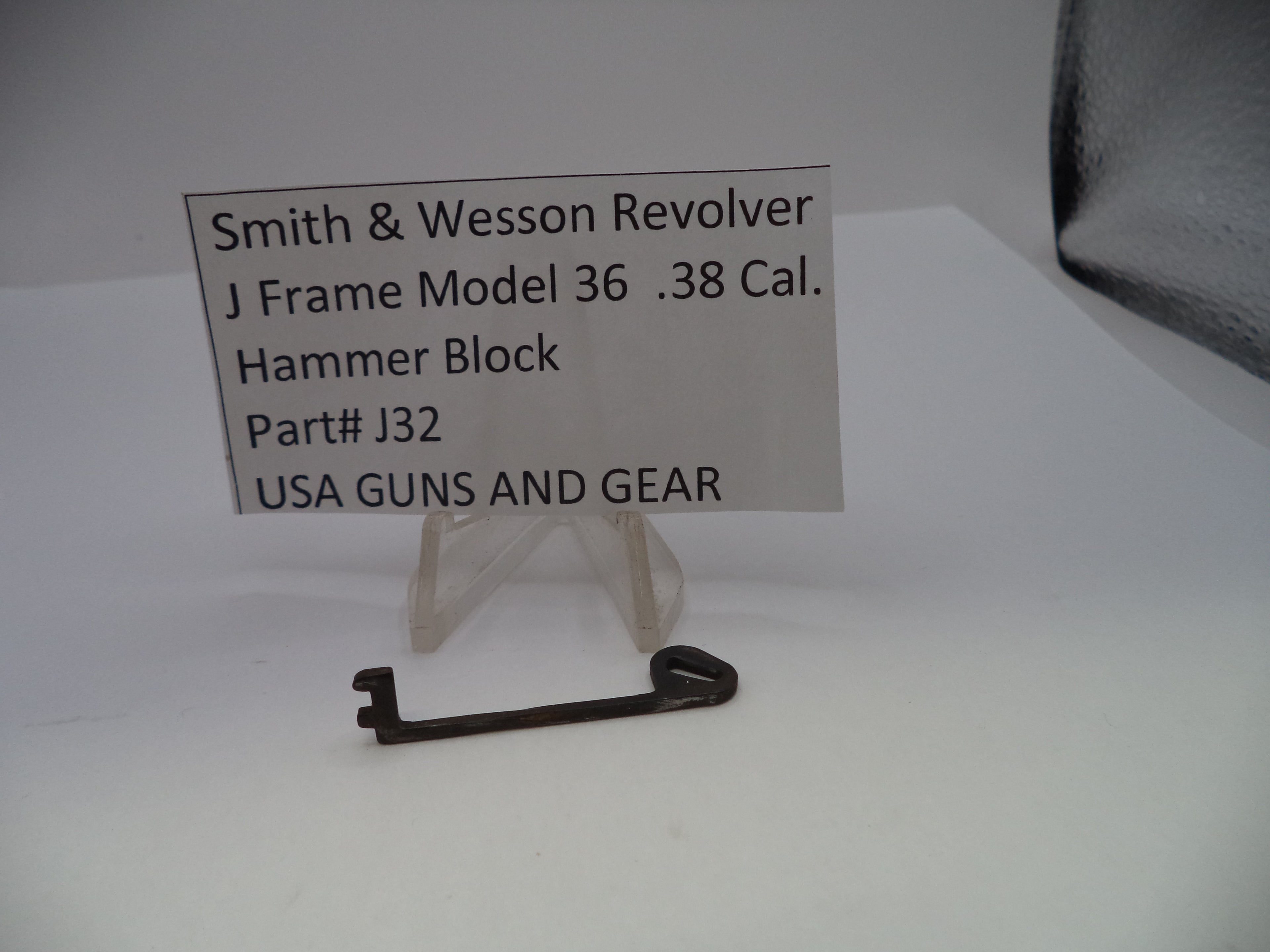 A small dark metal hammer block for a Smith & Wesson revolver J frame, displayed on a clear plastic stand next to a label card with product details.