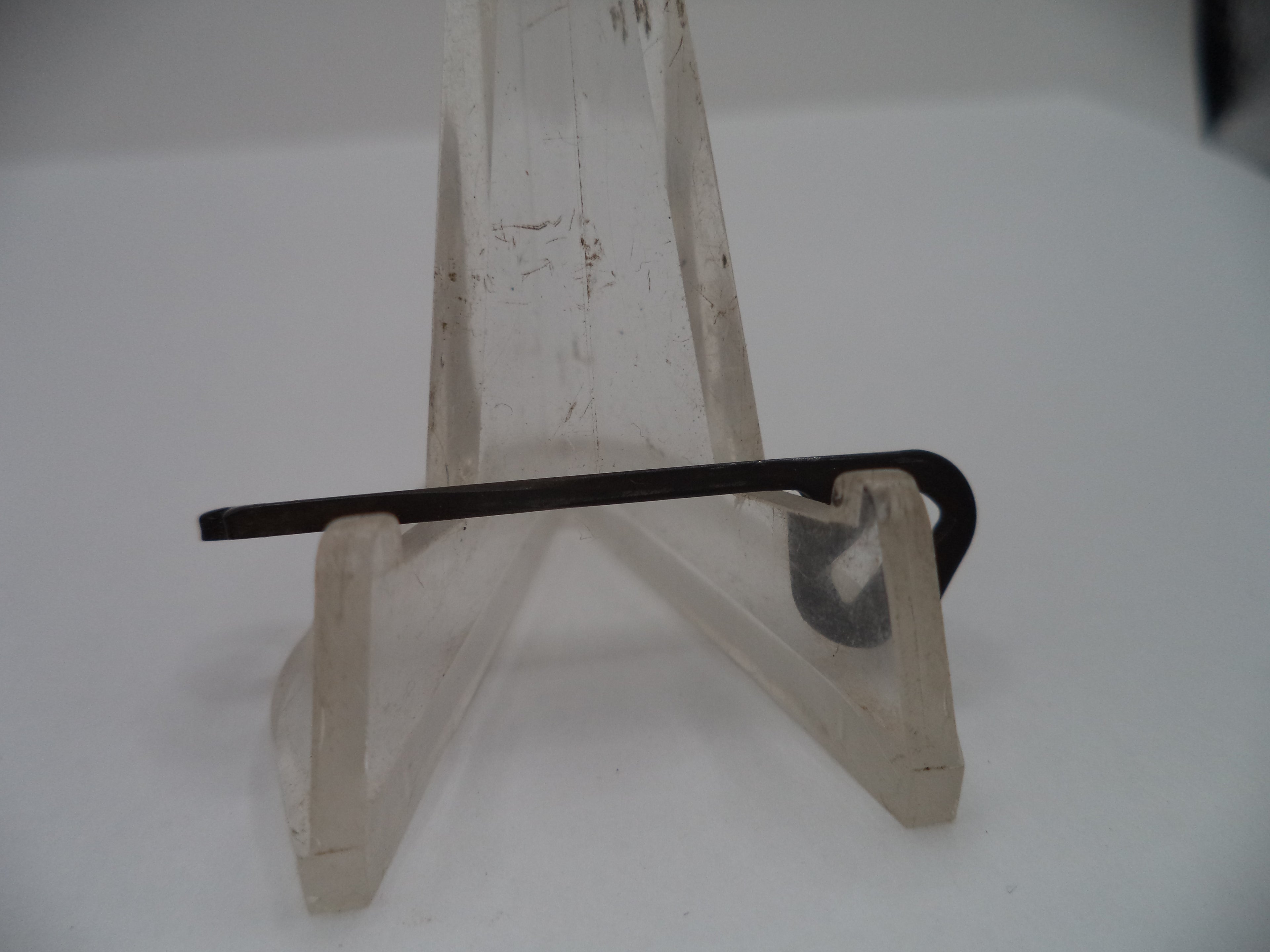 A small black metal tool with a curved hook or loop at one end, displayed on a clear plastic stand against a plain white background.