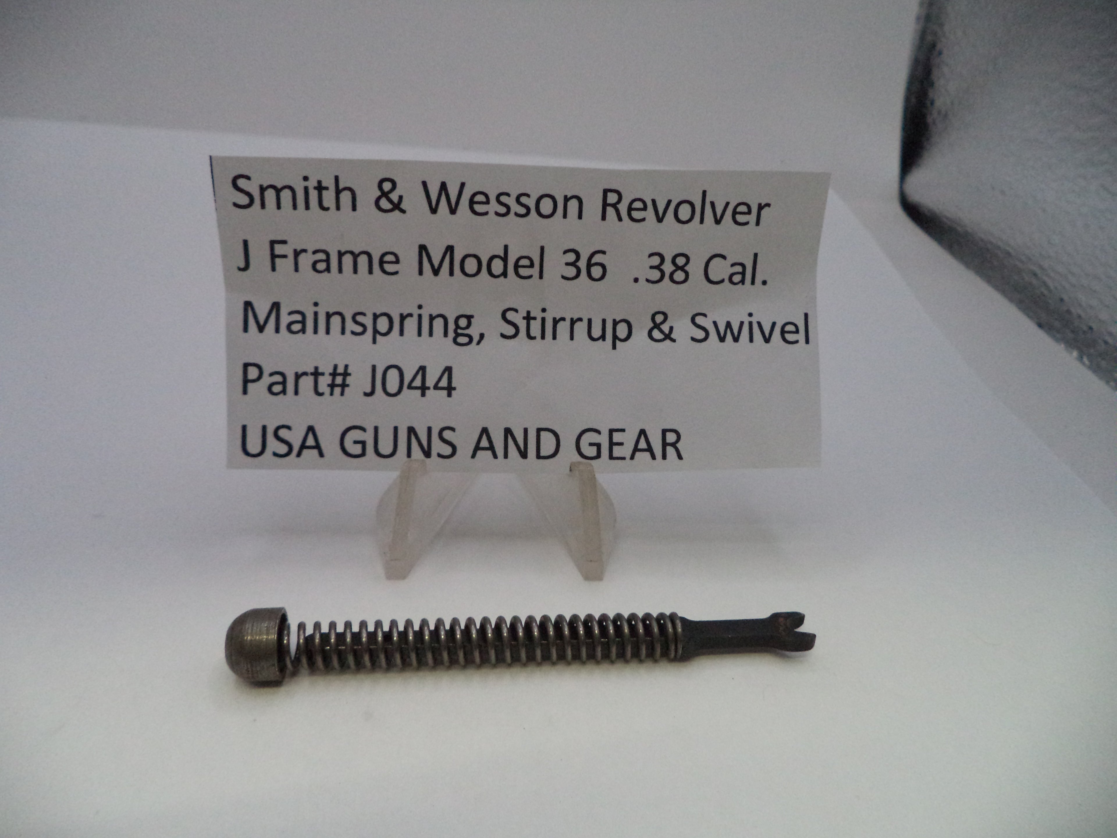A small metal revolver mainspring part displayed on a white surface with a small sign reading 'Smith & Wesson Revolver J Frame Model 36 .38 Cal.