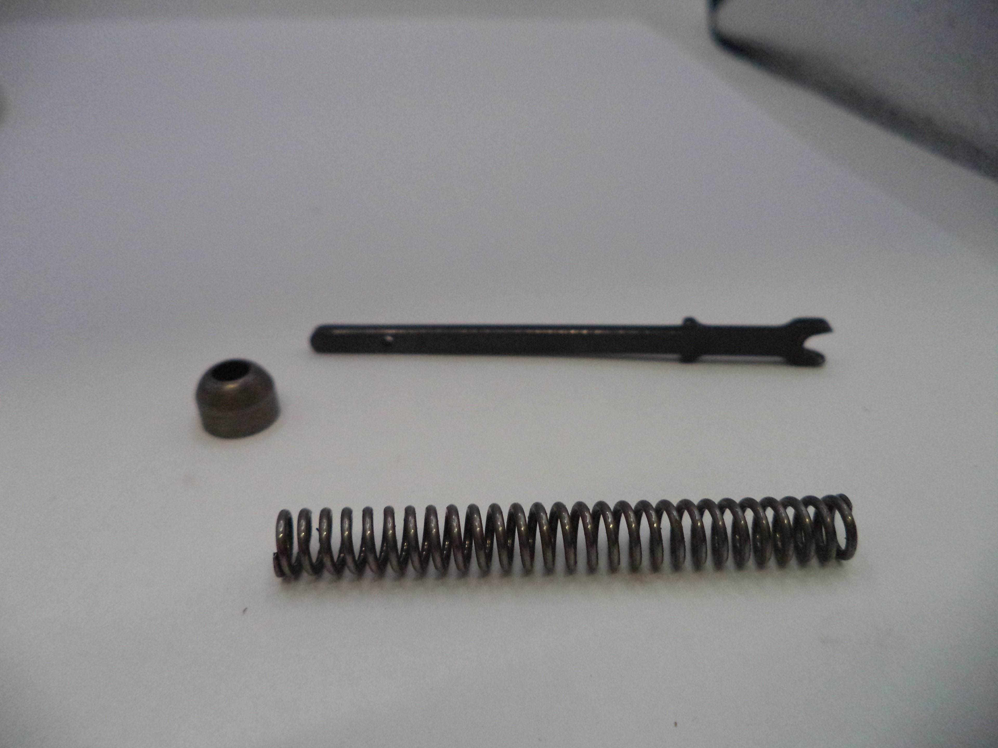 A small metal mechanical repair part set consisting of a long black metal pin with a bent tip, a small dark metal cap, and a coiled metal spring, arranged on a plain white surface.