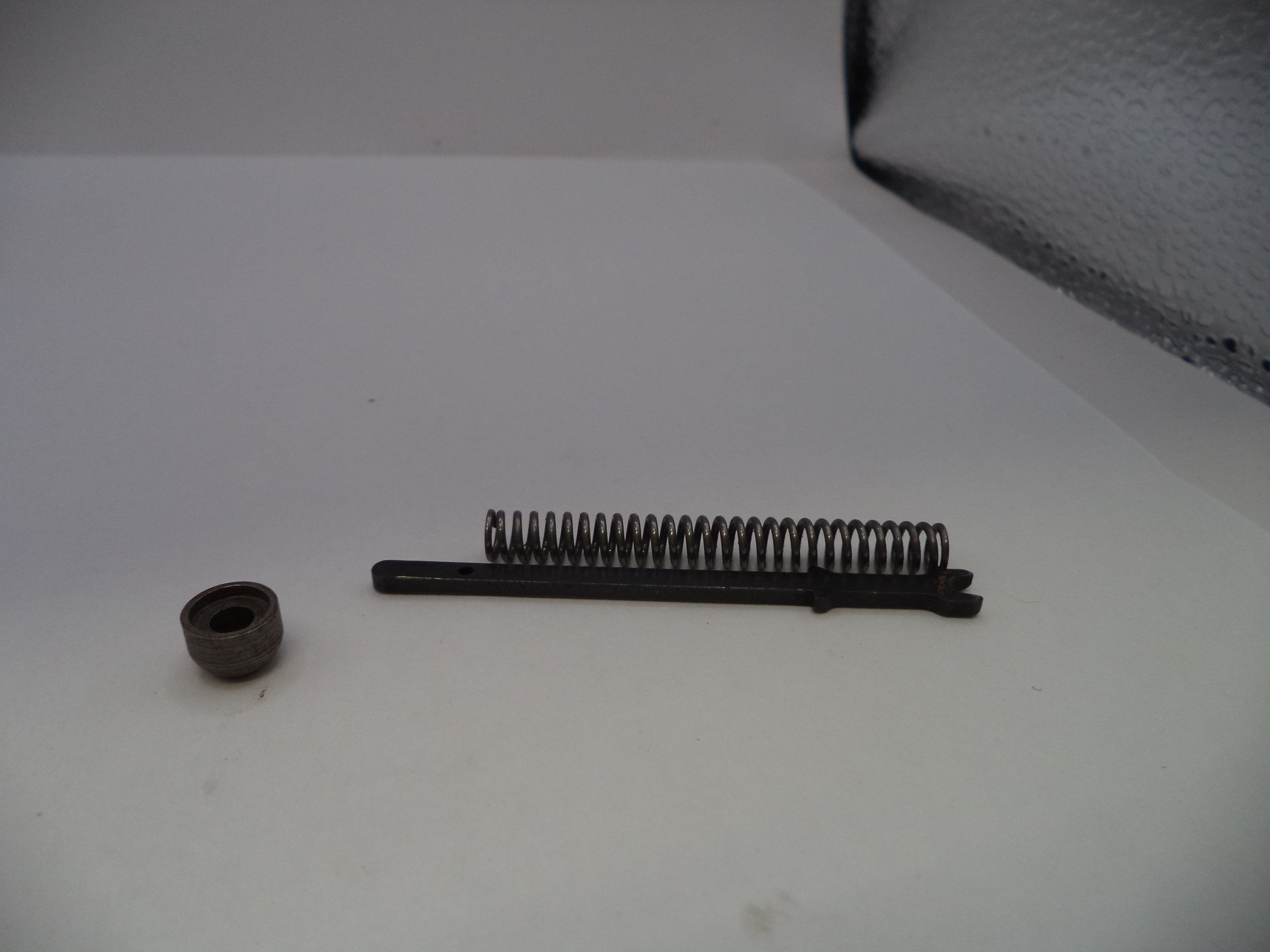 A small metal mechanical component set on a white surface, featuring a dark metal spring and a separate small circular piece.