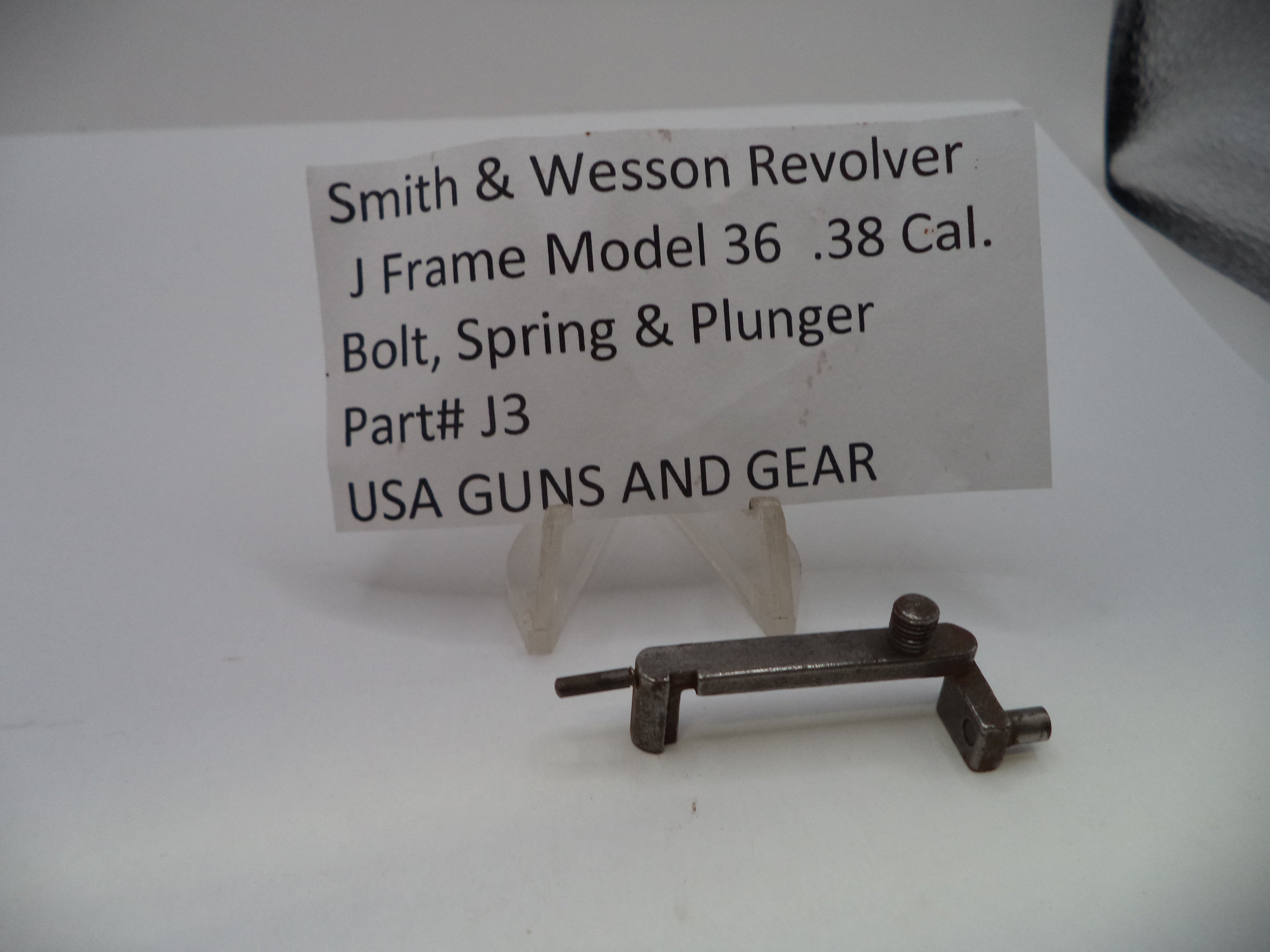 A small metal firearm part displayed on a clear plastic stand in front of a white label card that reads 'Smith & Wesson Revolver J Frame Model 36 .38 Cal.