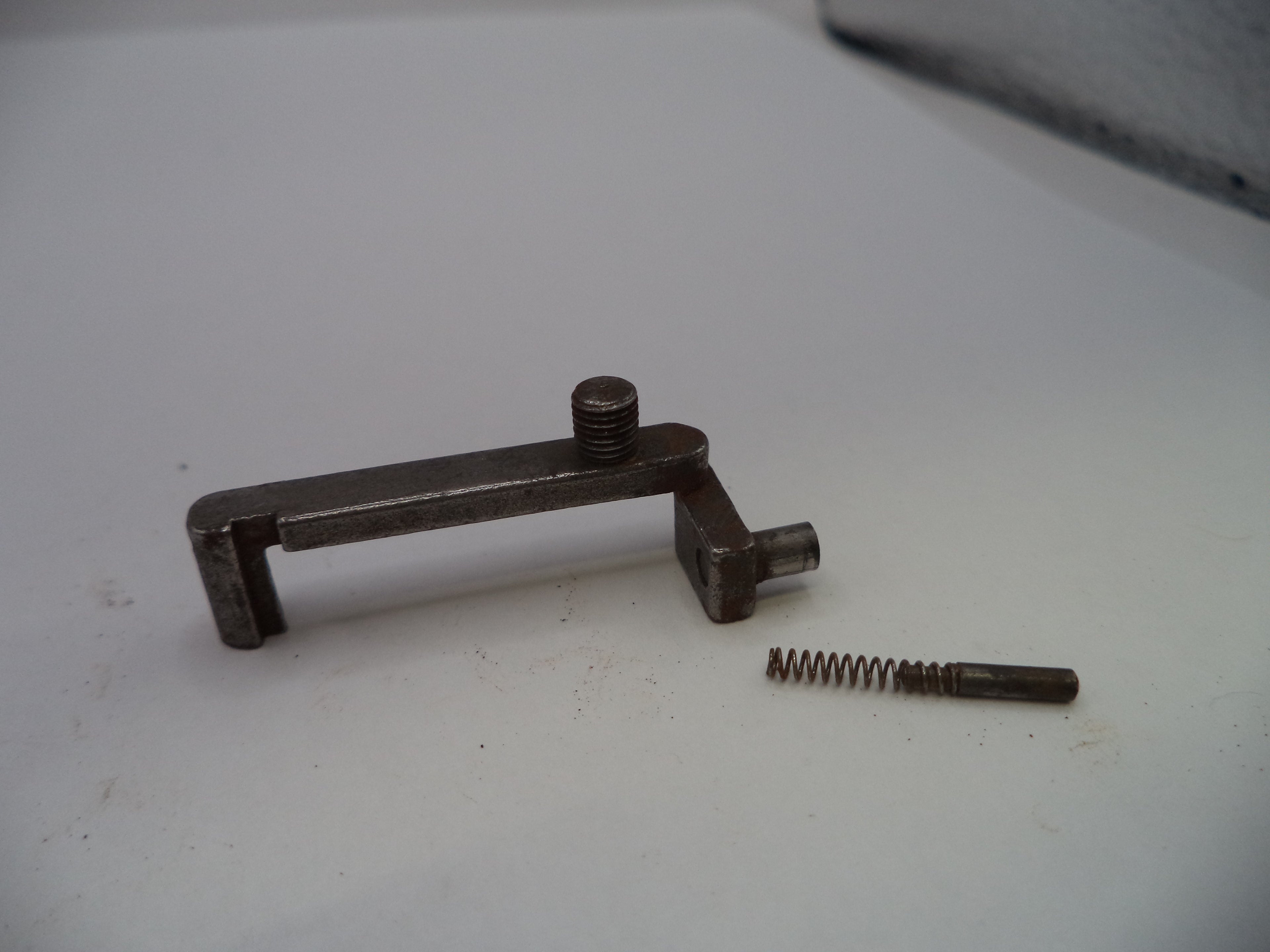 A small metal mechanical component with a screw and a separate coiled spring, photographed on a plain light surface.