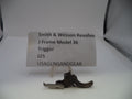 A small metal Smith & Wesson revolver trigger part displayed on a white surface with a paper label card behind it reading 'Smith & Wesson Revolver J Frame Model 36 Trigger J25 USAGUNSANDGEAR'.