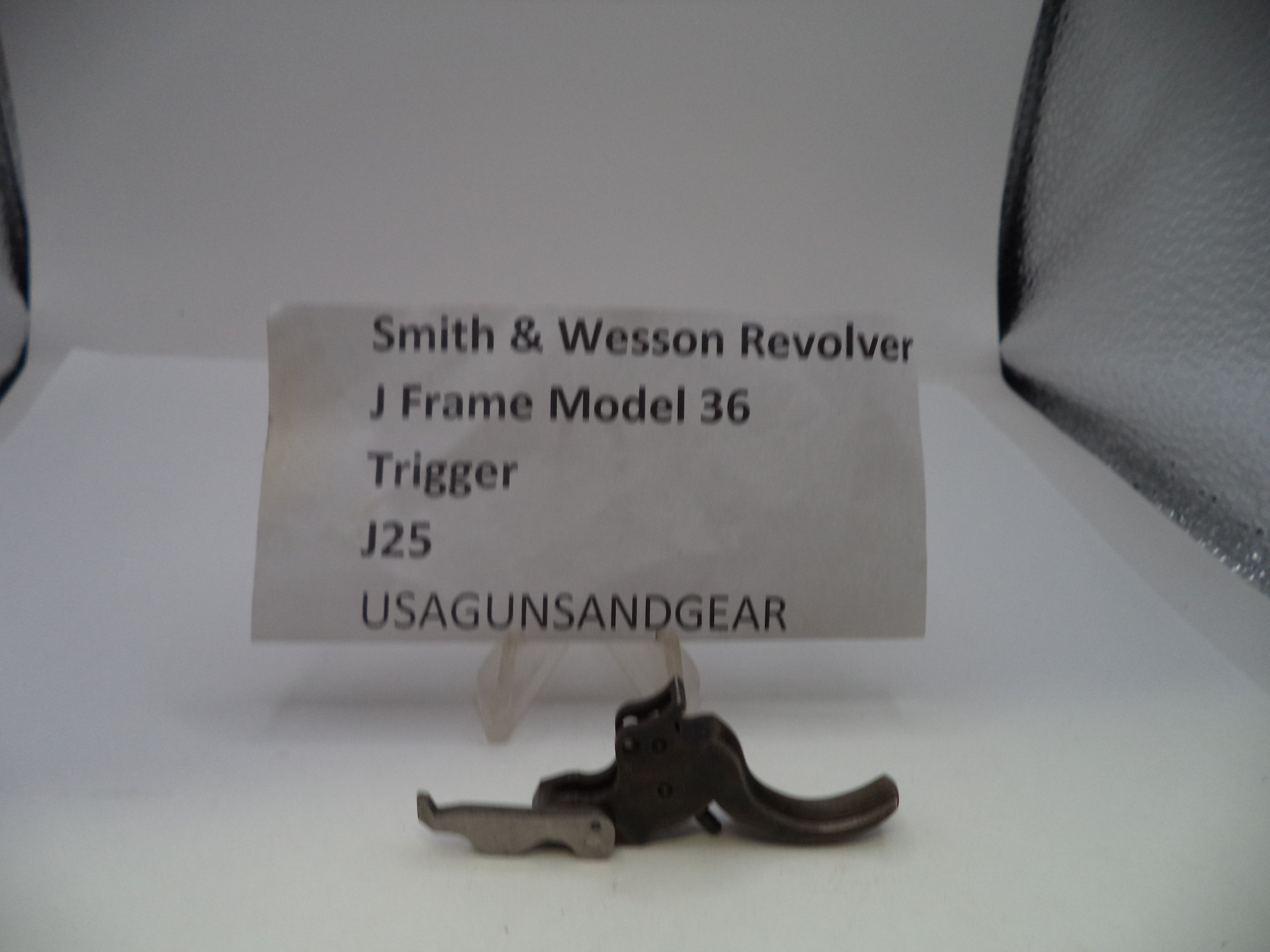 A small metal Smith & Wesson revolver trigger part displayed on a white surface with a paper label card behind it reading 'Smith & Wesson Revolver J Frame Model 36 Trigger J25 USAGUNSANDGEAR'.