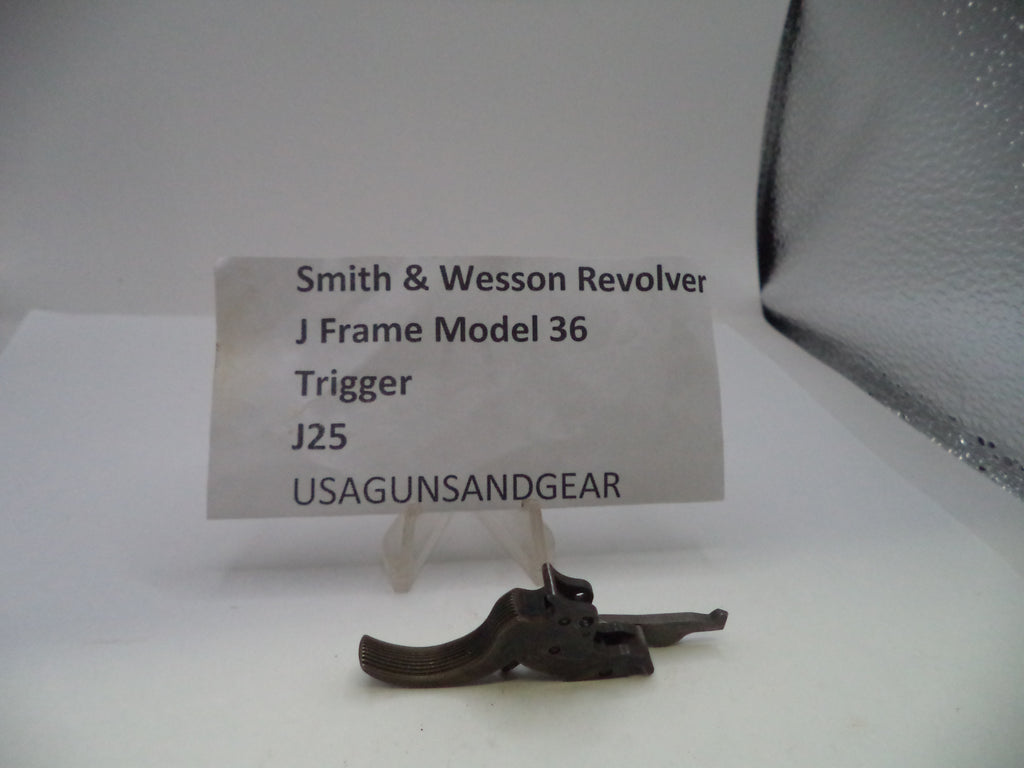 A small metal revolver trigger part displayed on a clear stand in front of a white label card that reads 'Smith & Wesson Revolver J Frame Model 36 Trigger J25 USAGUNSANDGEAR'.
