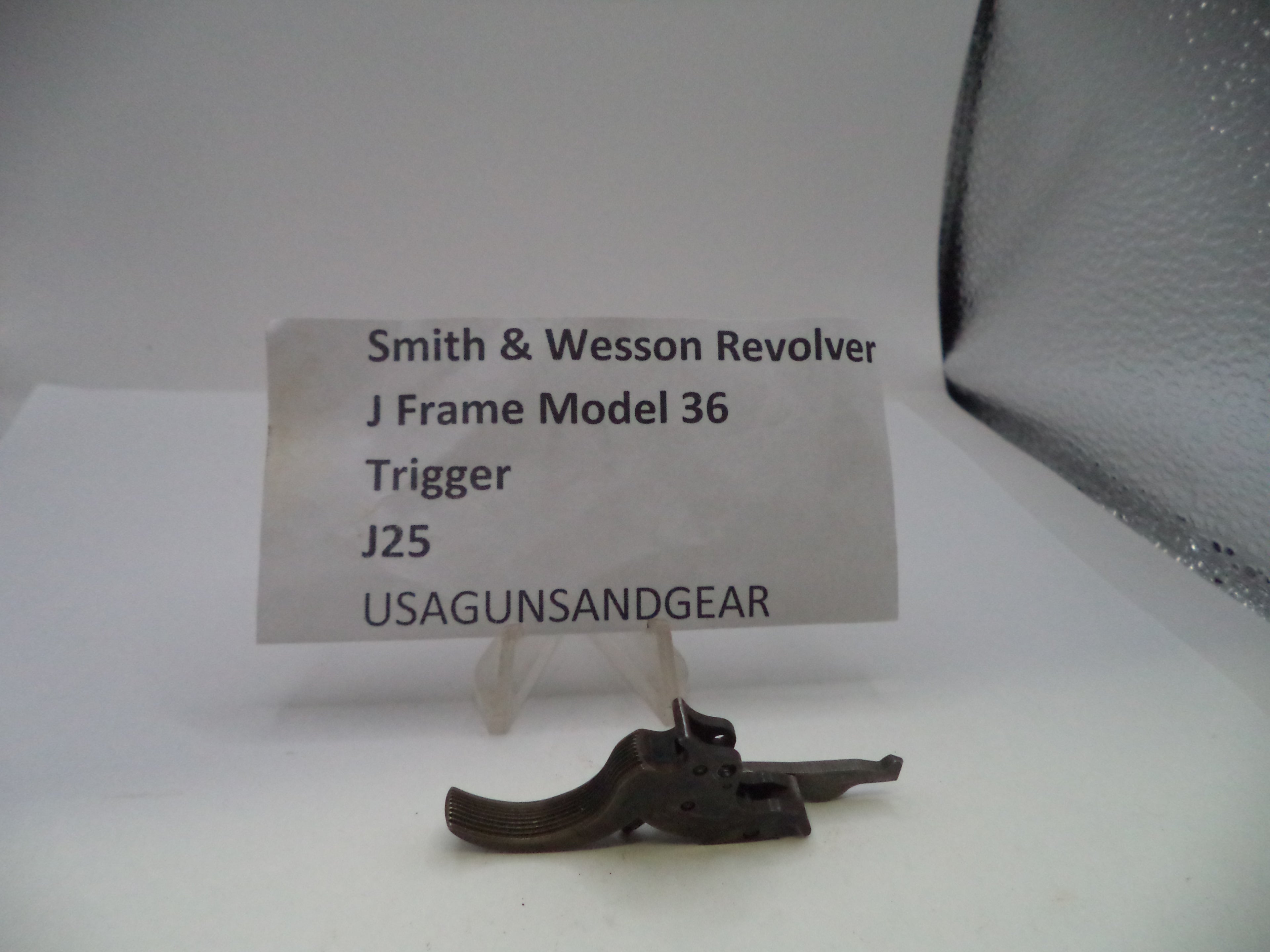 A small metal revolver trigger part displayed on a clear stand in front of a white label card that reads 'Smith & Wesson Revolver J Frame Model 36 Trigger J25 USAGUNSANDGEAR'.