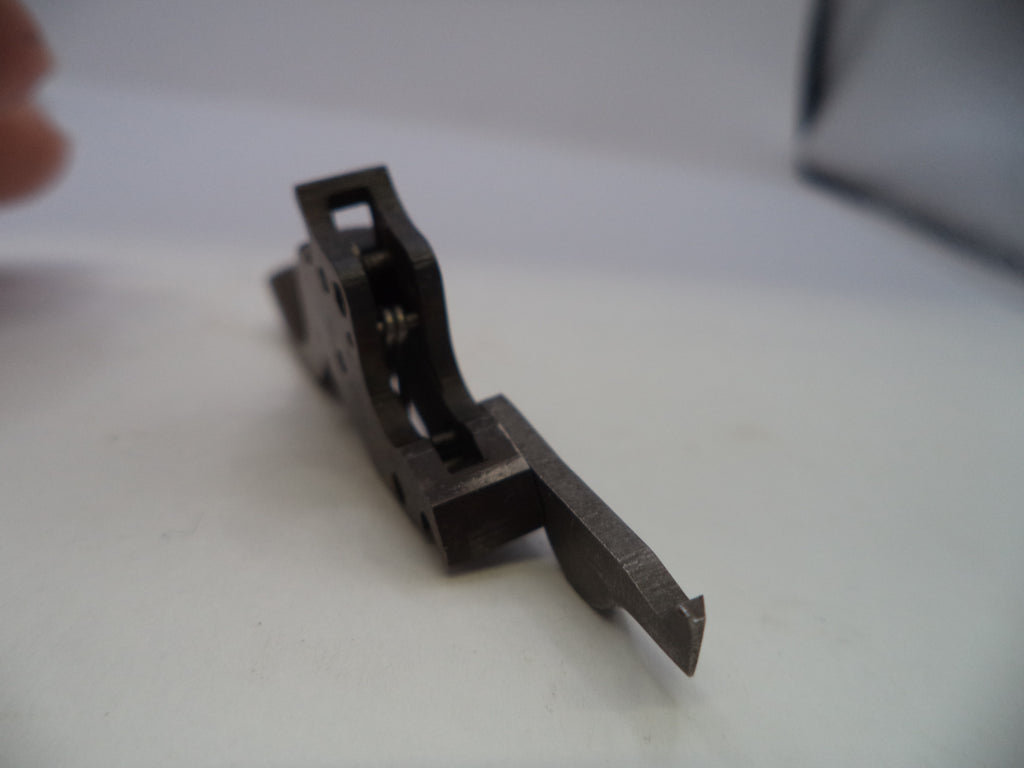 A small dark metal mechanical component or tool part photographed on a plain light surface, with a finger partially visible at the upper left and a blurred object at the upper right.