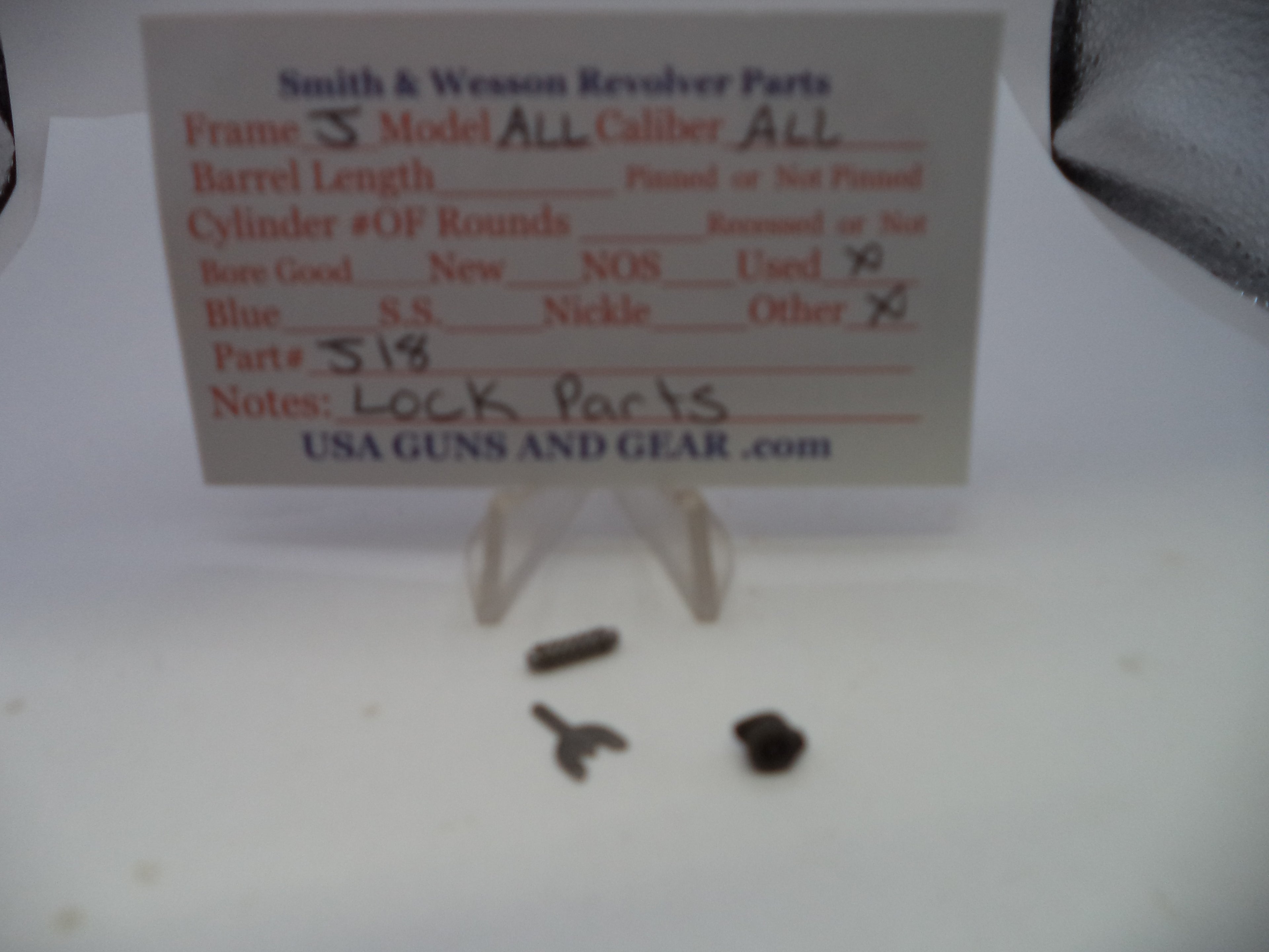 Small metal revolver lock parts displayed on a clear stand in front of a handwritten parts card labeled Smith & Wesson Revolver Parts.