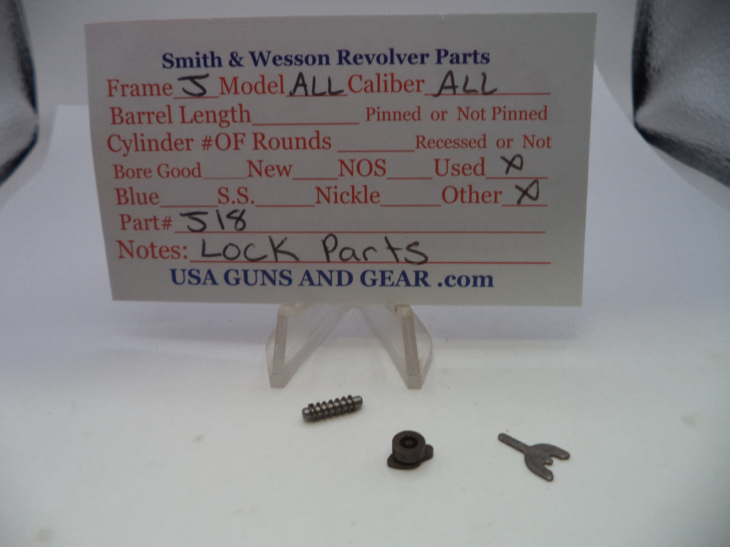 Smith & Wesson revolver lock parts displayed on a white surface with a handwritten parts card. The set includes a small metal screw, a dark metal lock component, and a small metal key, shown on clear plastic stands.