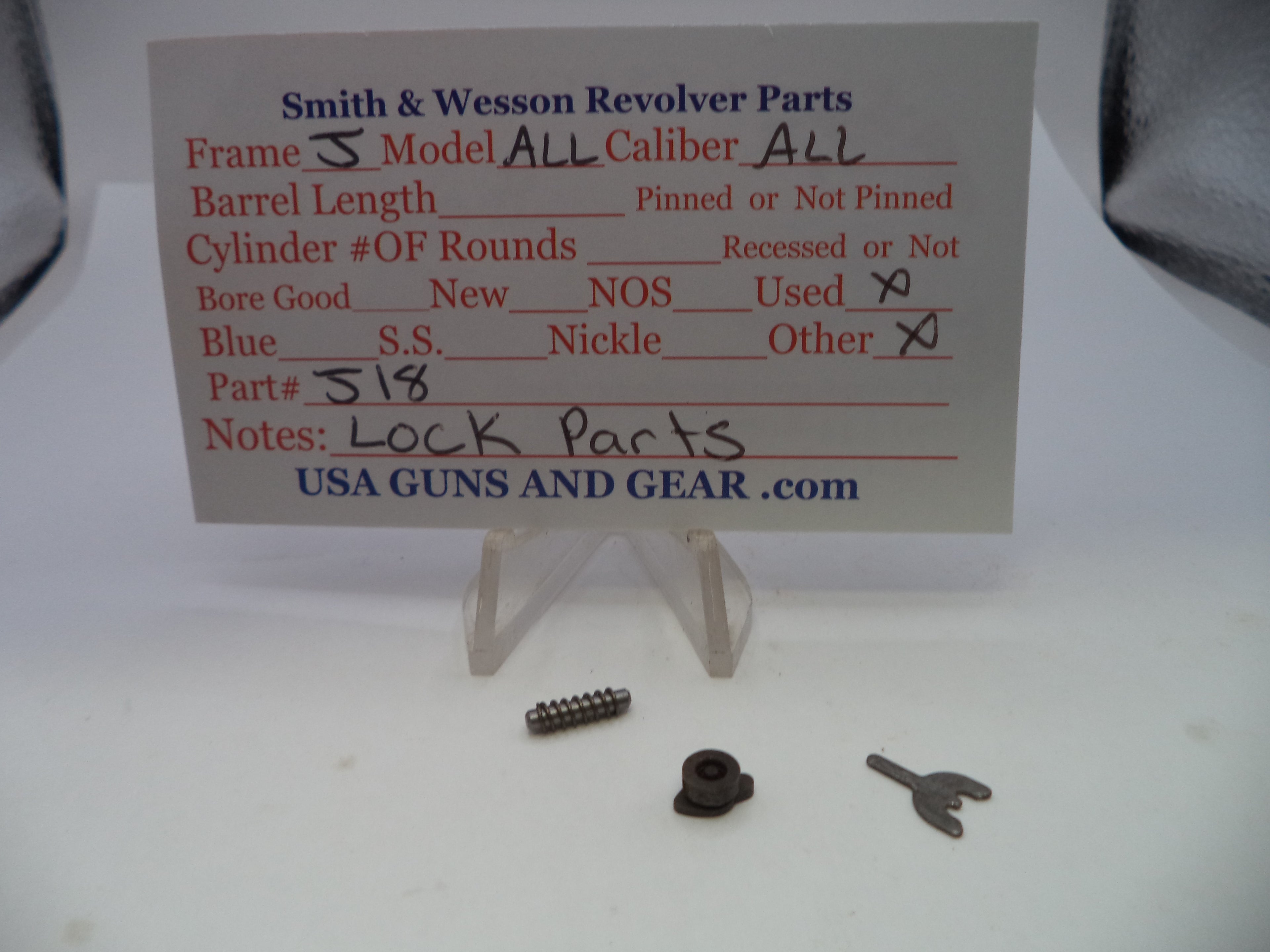 Smith & Wesson revolver lock parts displayed on a white surface with a handwritten parts card. The set includes a small metal screw, a dark metal lock component, and a small metal key, shown on clear plastic stands.