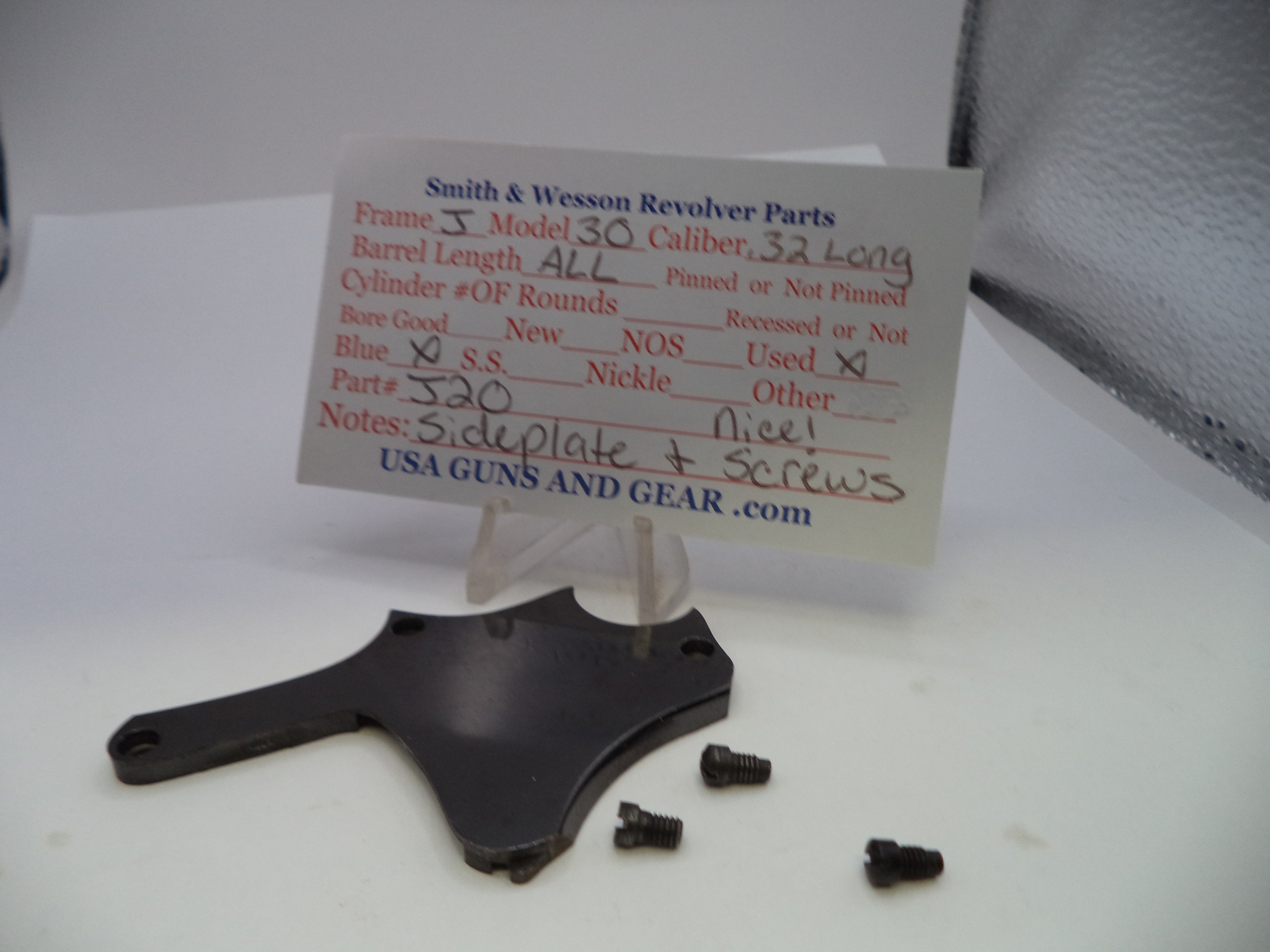 A black metal revolver part with three small black screws displayed on a white surface, with a handwritten paper label card behind it reading Smith & Wesson Revolver Parts.