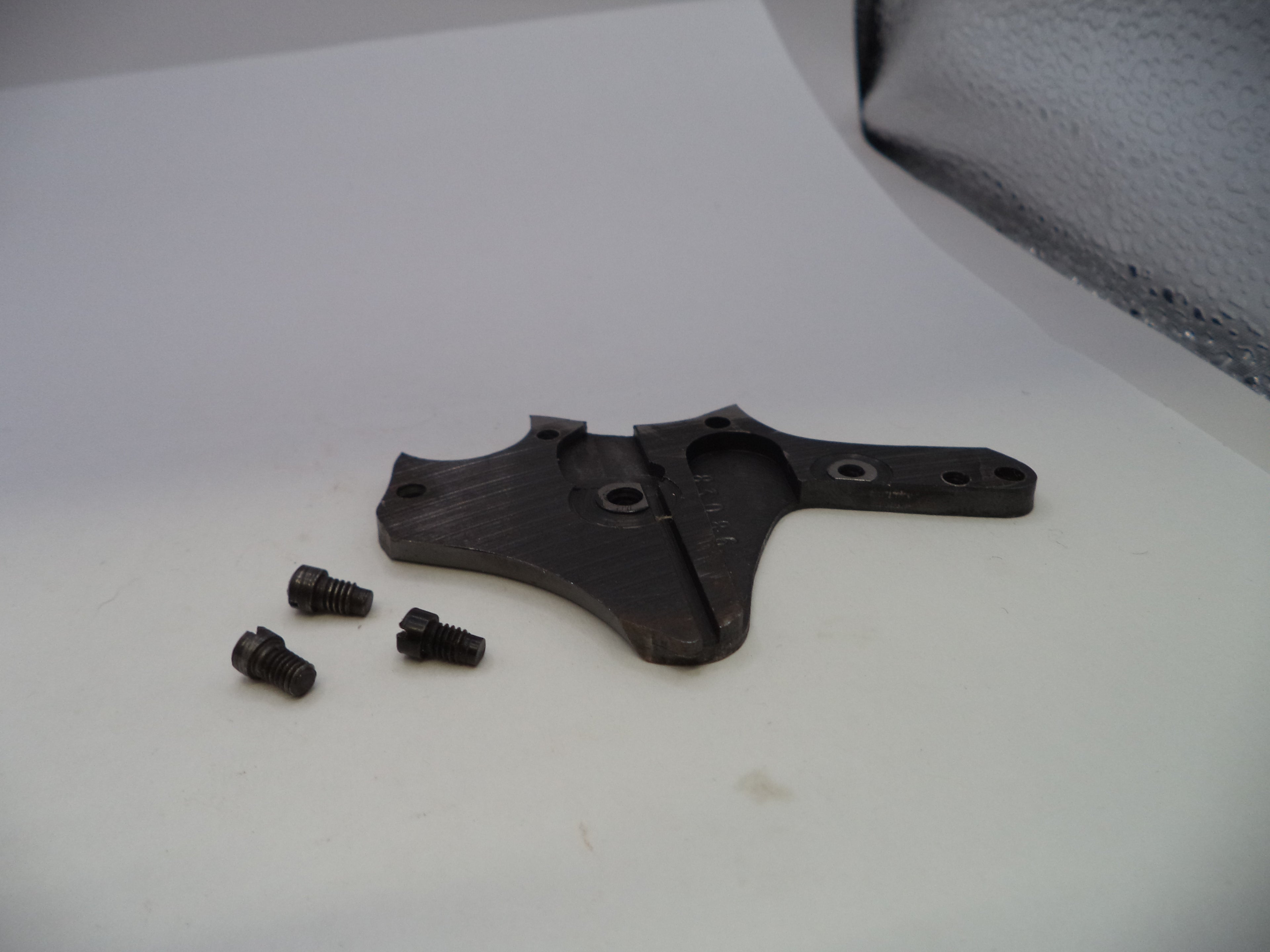 A black metal mechanical bracket or plate with three small black screws placed beside it on a white surface.