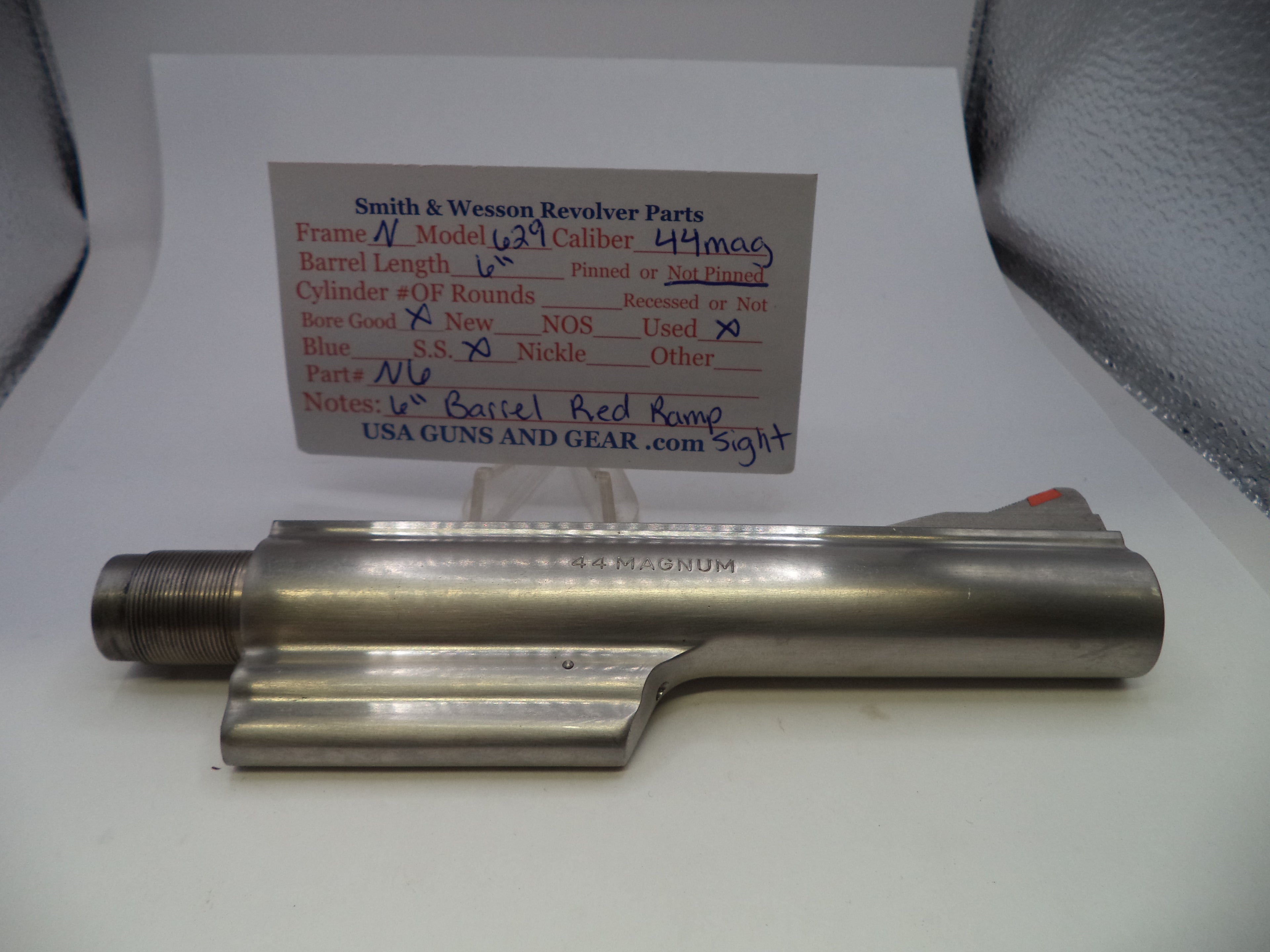 A metallic cylindrical gun part with a threaded end and a flat side, photographed on a white surface with a handwritten parts card behind it. The card reads Smith & Wesson Revolver Parts and includes caliber and part details.