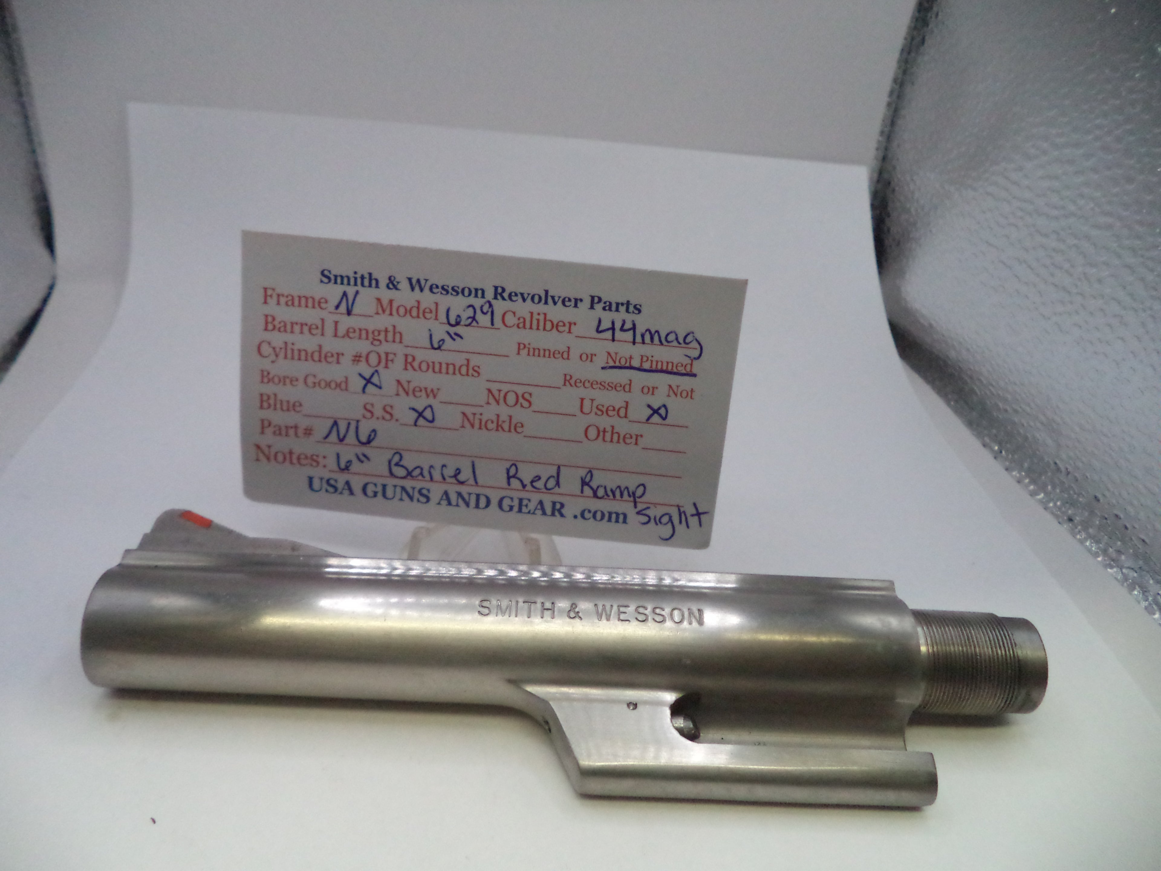 A Smith & Wesson revolver cylinder block part made of stainless steel, displayed on a white surface with a handwritten parts identification card behind it.