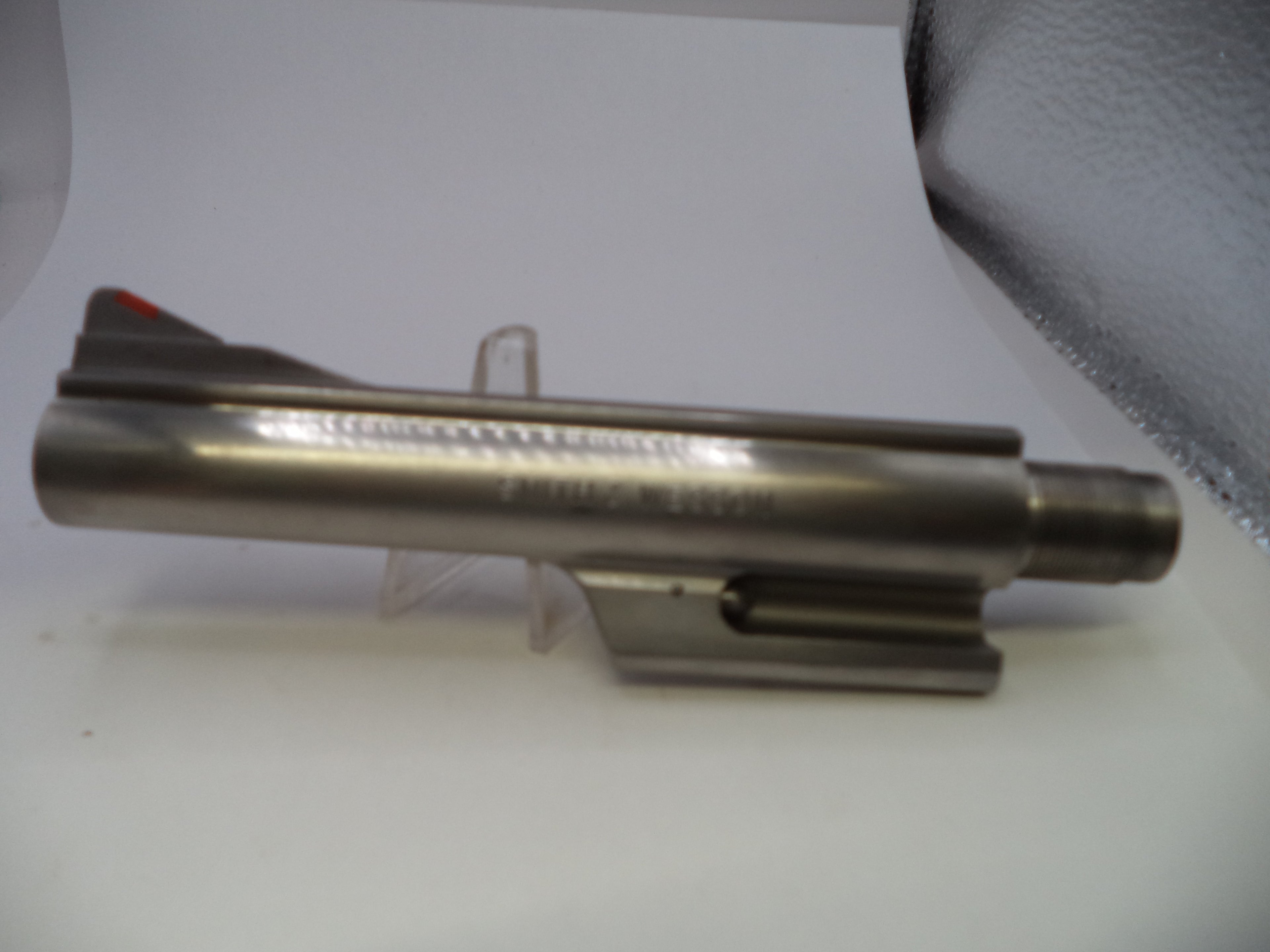 A metallic gun barrel or slide component displayed on a small clear stand against a white background with a textured gray area in the upper right.