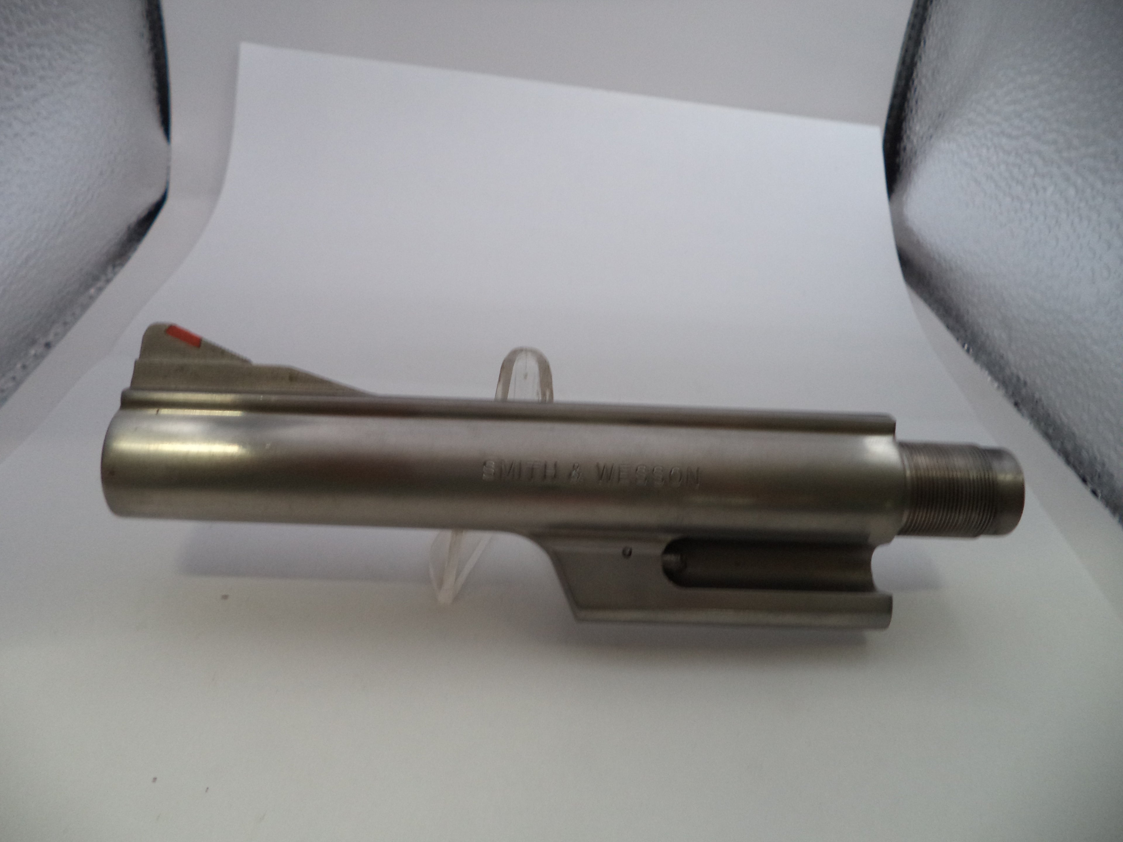A metallic gun barrel or receiver component displayed on a clear stand against a white background with gray textured side panels.