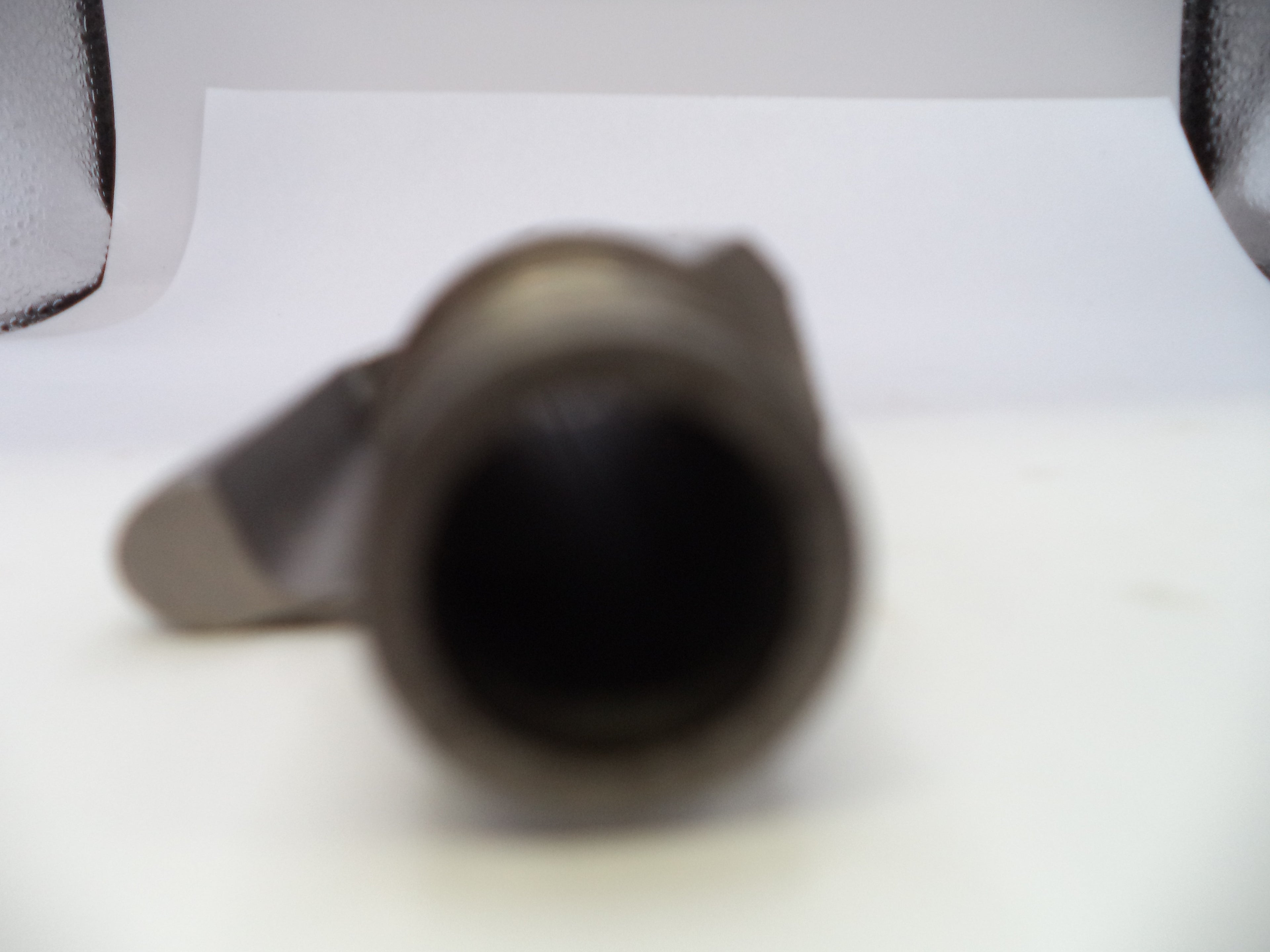 A close-up, heavily blurred shot of a dark metallic cylindrical object, possibly a mechanical part or tool, resting on a white surface.
