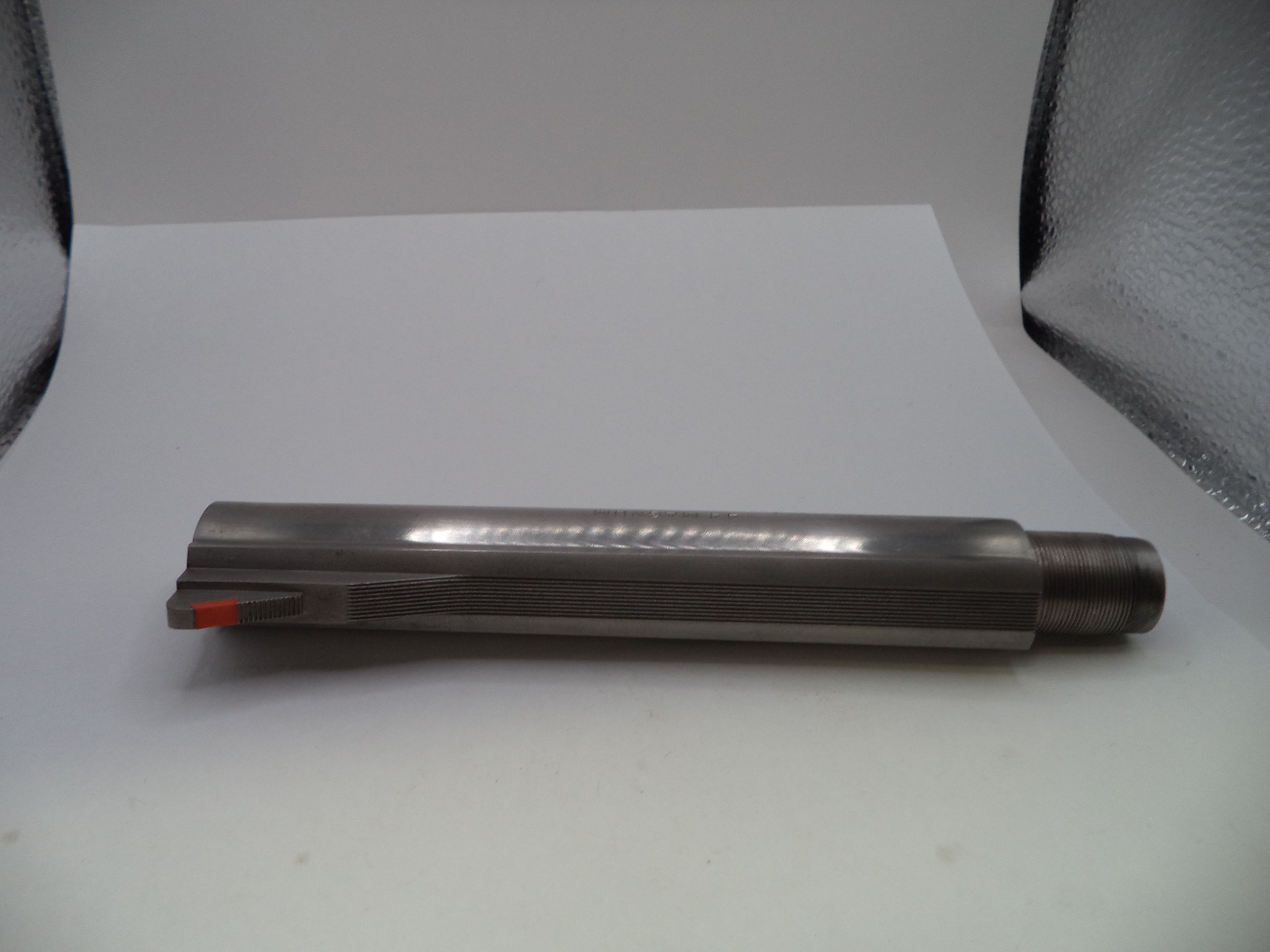 A cylindrical metal cutting tool or drill bit with a fluted shank and a small orange accent near the cutting edge, photographed on a white surface with a reflective silver backdrop.
