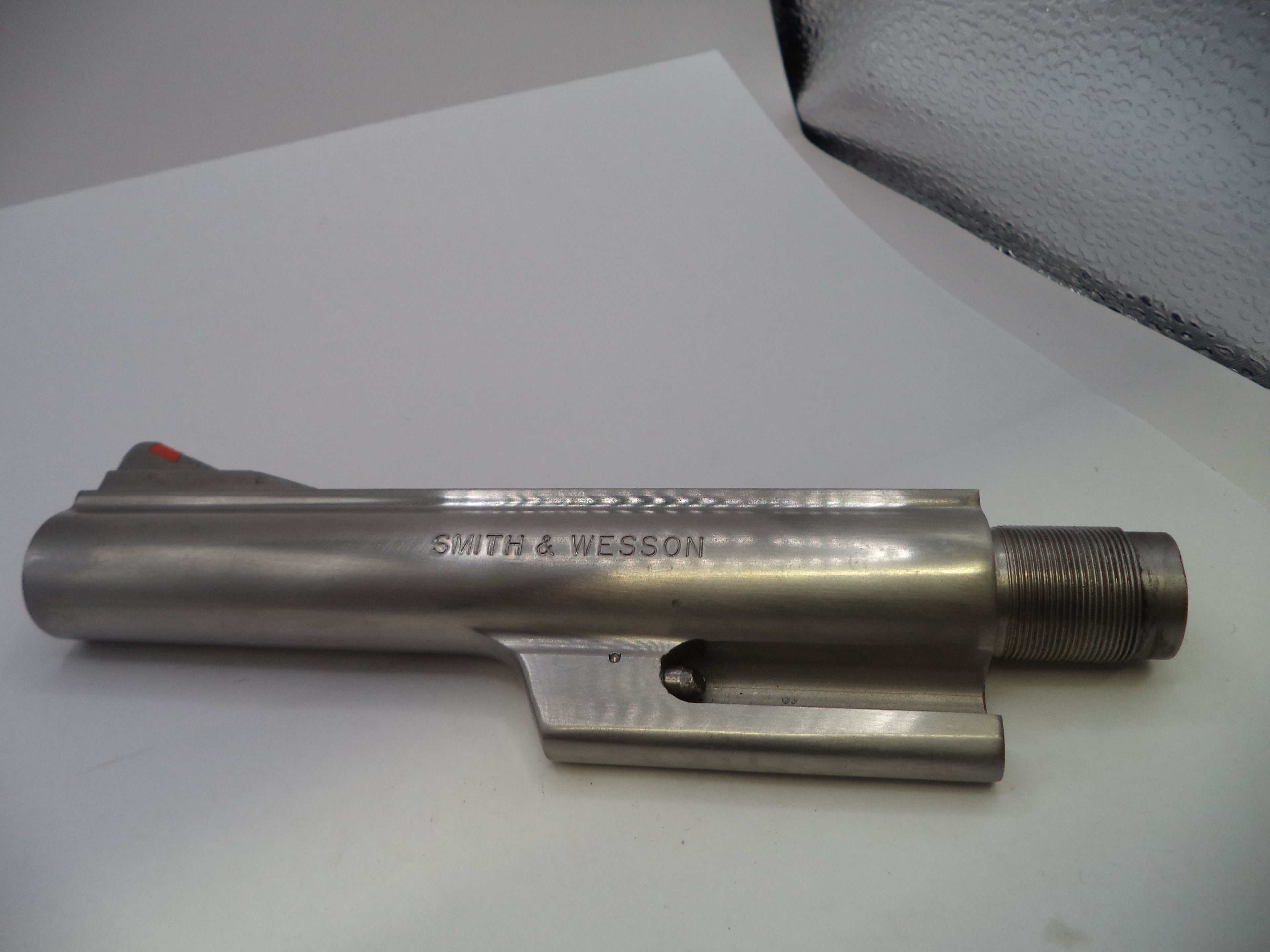 A metallic Smith & Wesson receiver or barrel component photographed on a white surface with a textured gray background visible in the upper right.