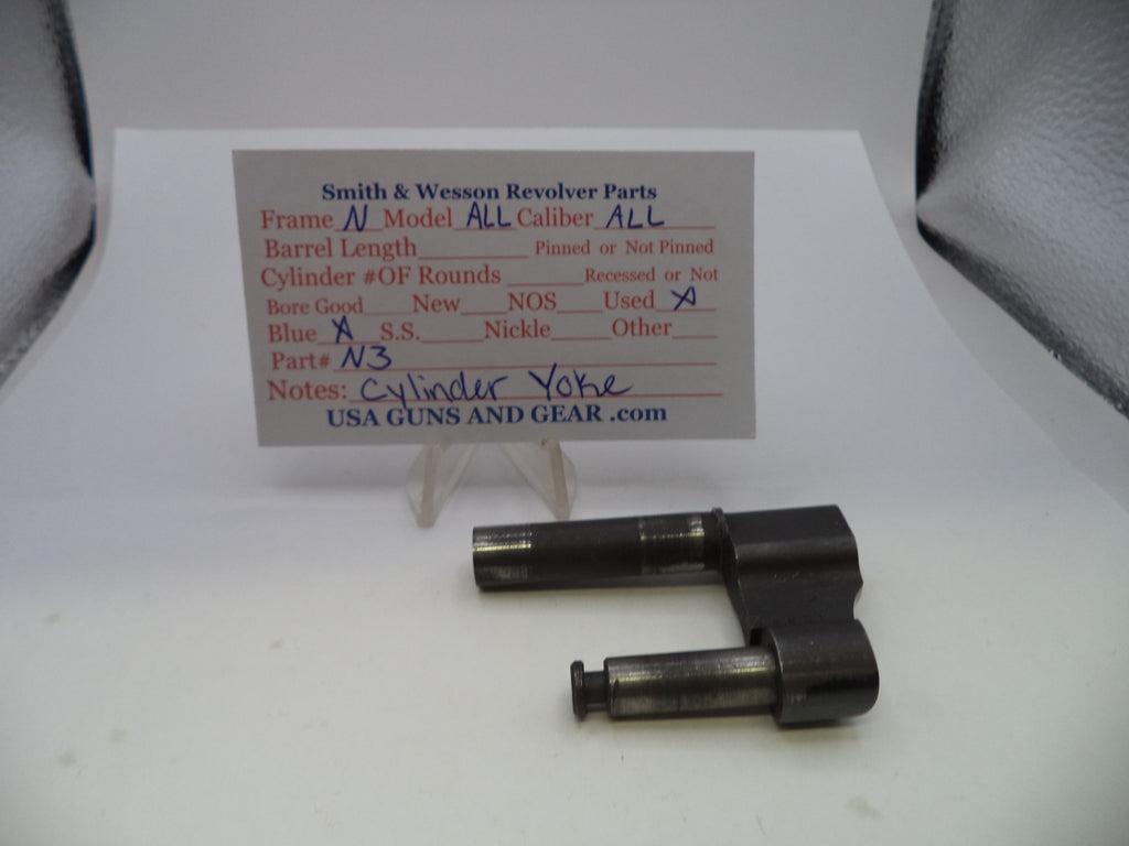 A used black metal revolver cylinder yoke part displayed on a white surface with a handwritten parts identification card behind it.