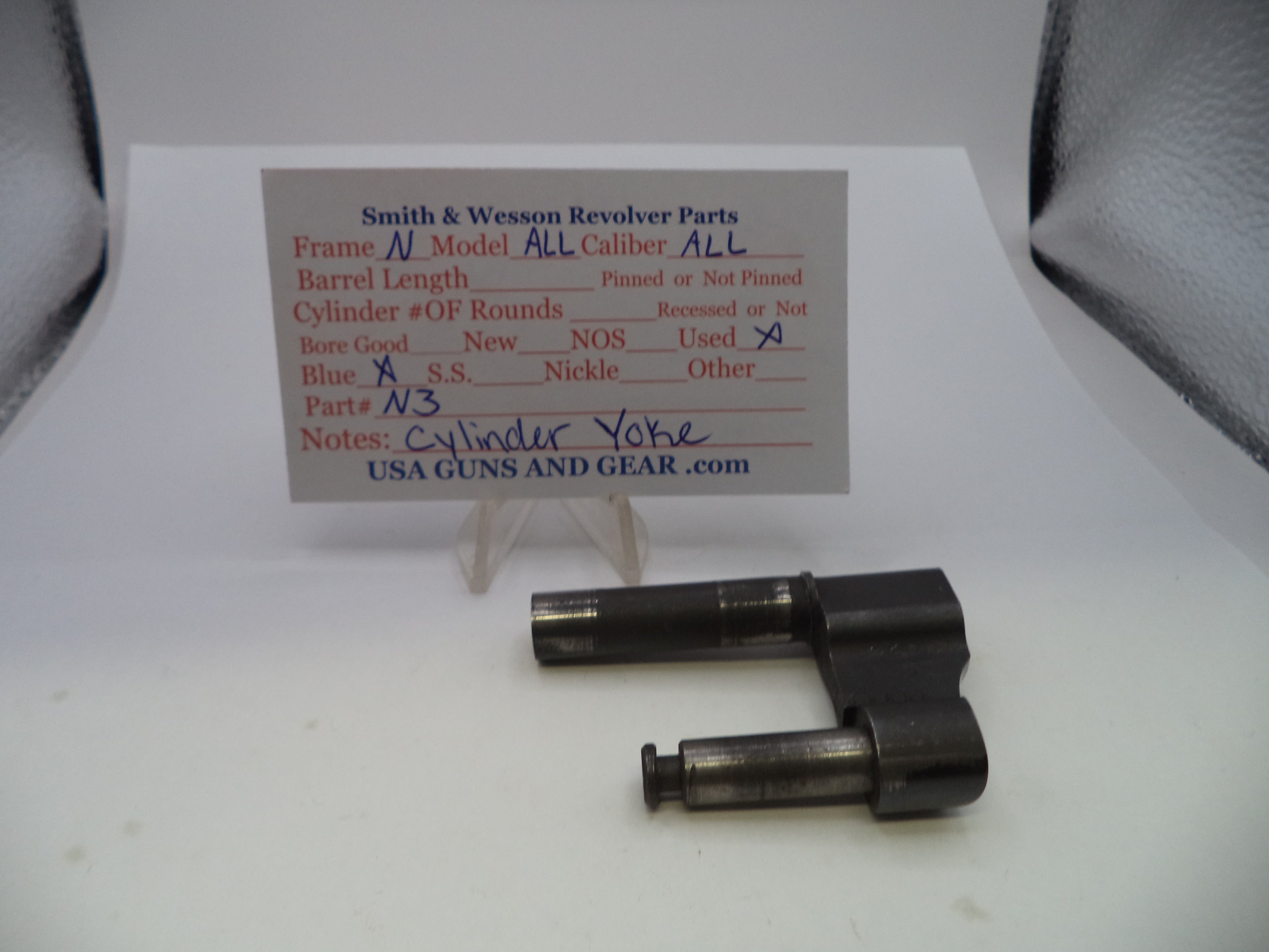 A used black metal revolver cylinder yoke part displayed on a white surface with a handwritten parts identification card behind it.