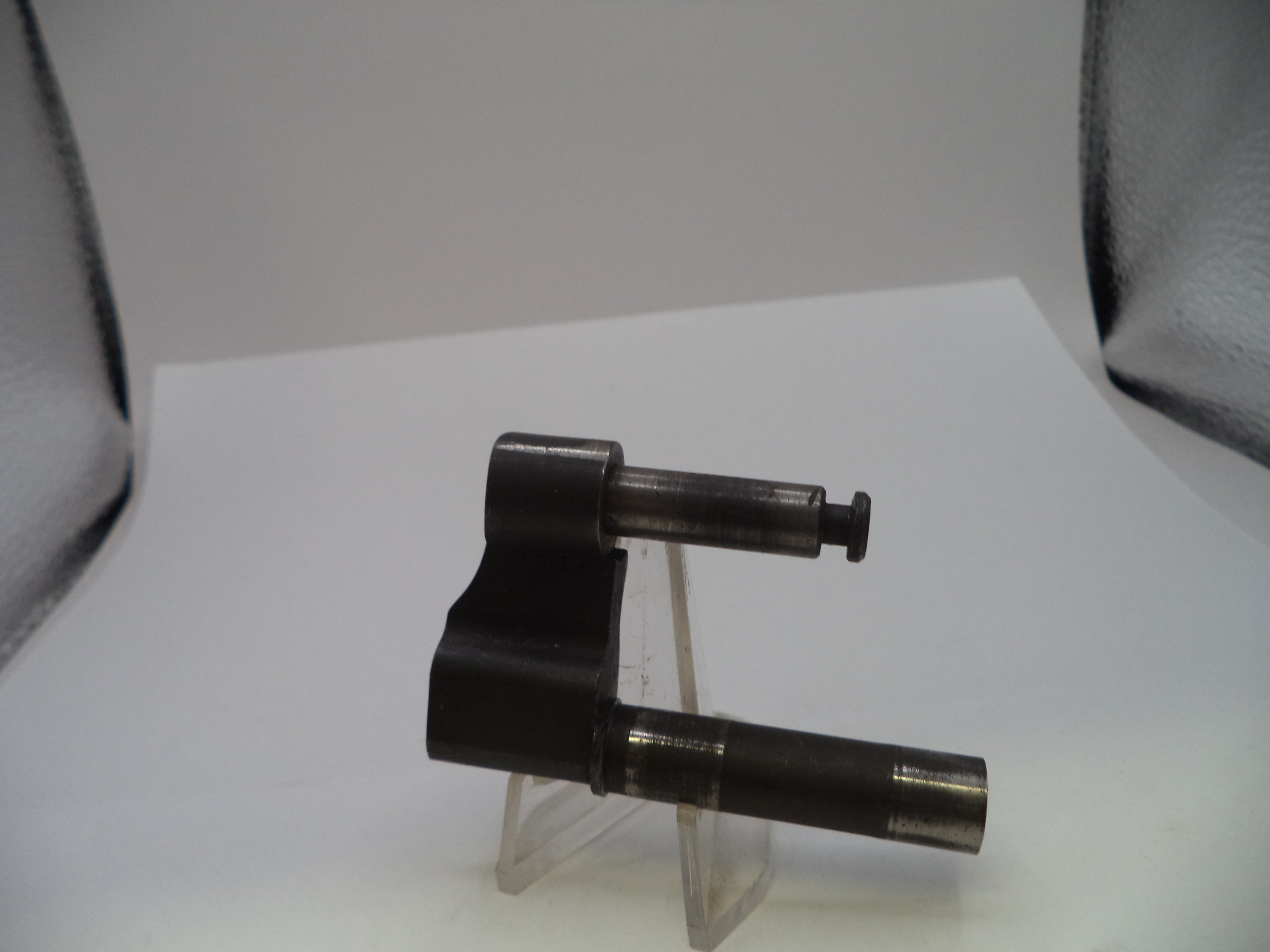 A dark metallic mechanical part, possibly a valve stem or throttle body component, displayed on a clear plastic stand against a white background with studio lights visible in the corners.