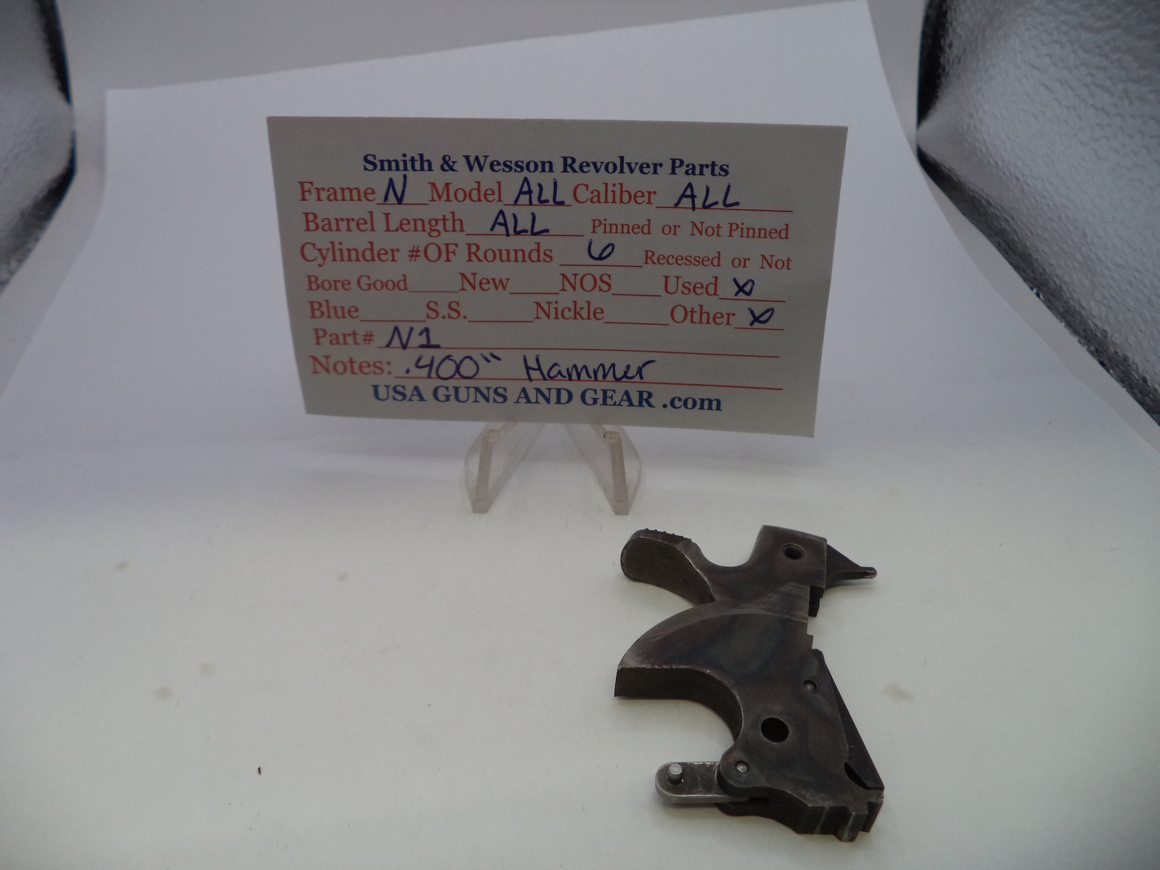 A dark metal revolver replacement part displayed on a white surface next to a handwritten parts identification card for a Smith & Wesson revolver.