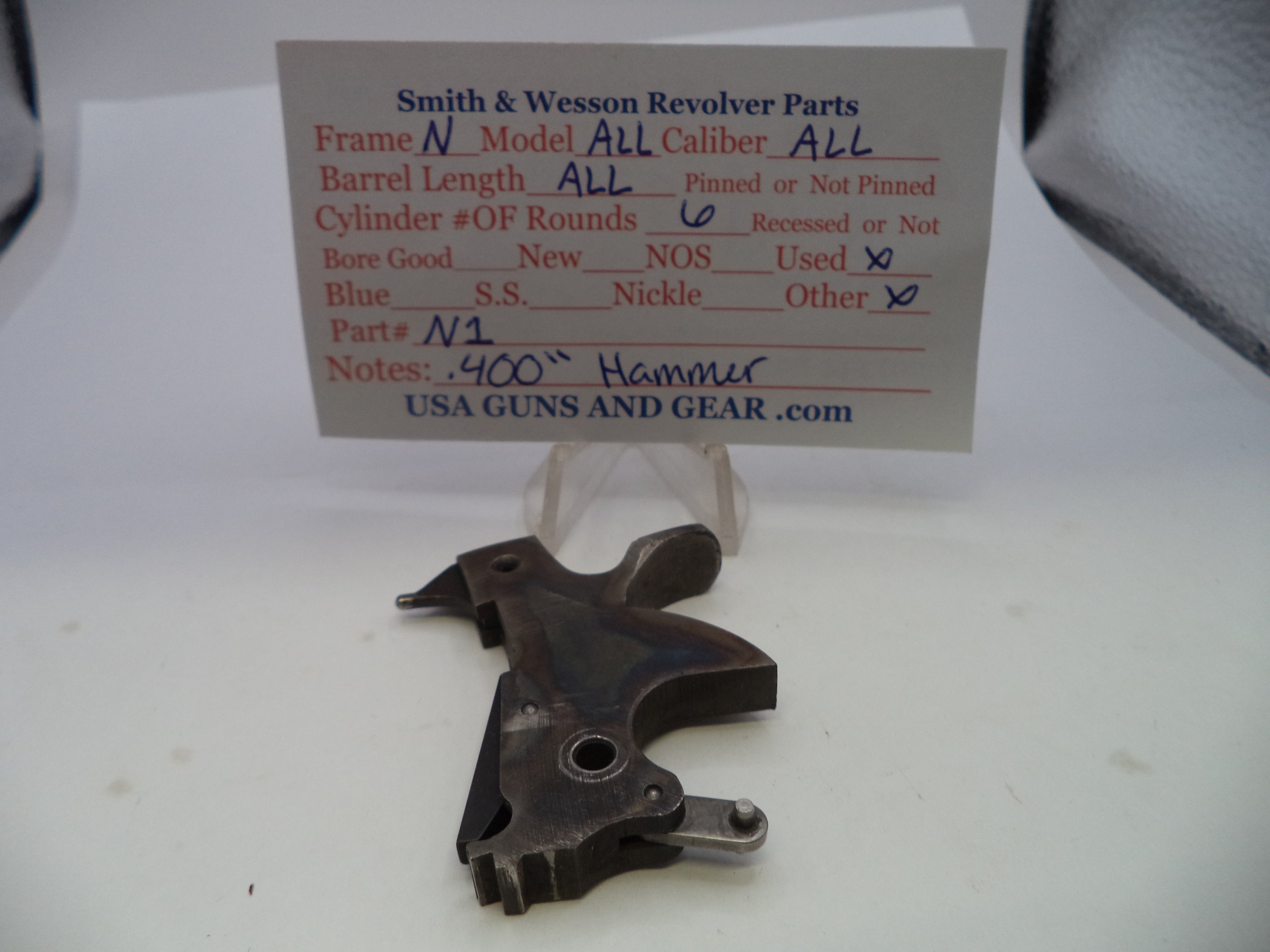 A used metal Smith & Wesson revolver part, likely a grip or frame component, displayed in front of a handwritten inventory card on a white surface.