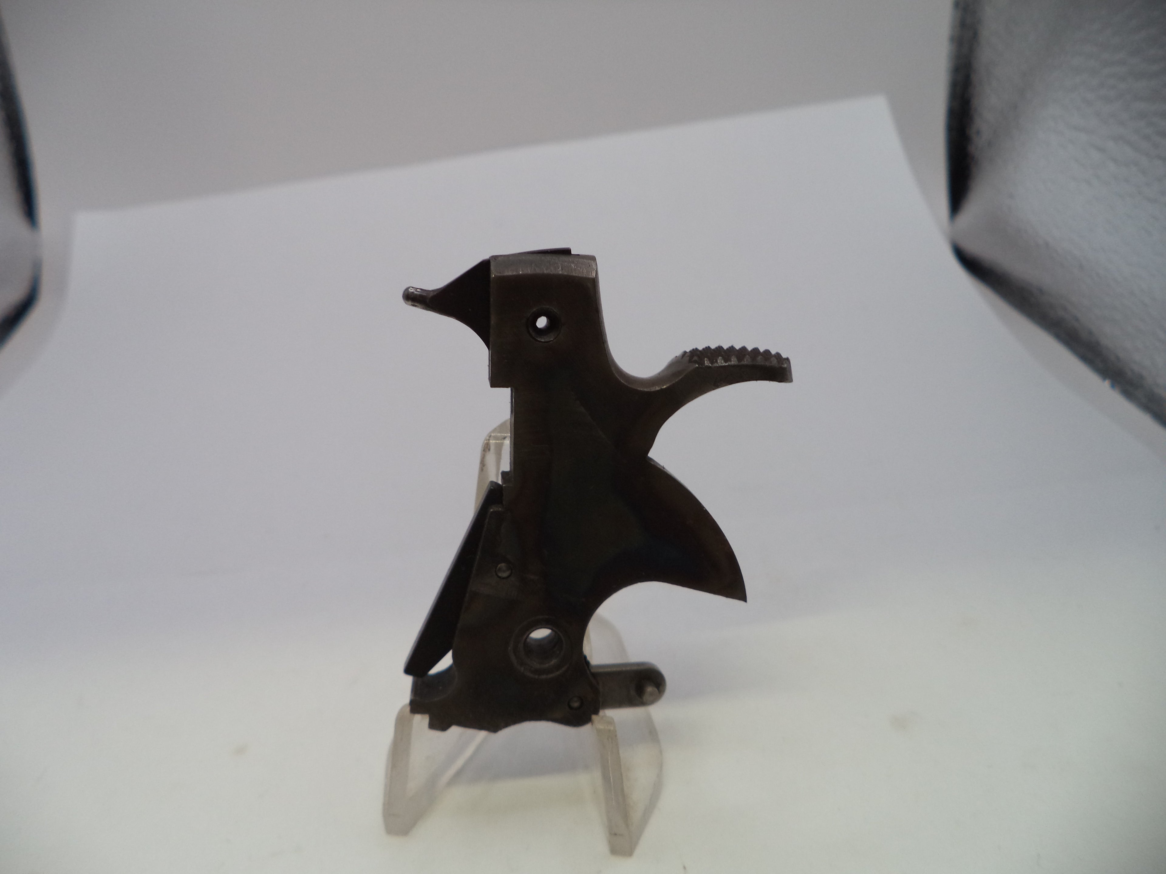 A dark metal trigger assembly for a firearm, displayed on a clear plastic stand against a white background with a black textured surface visible in the upper right.