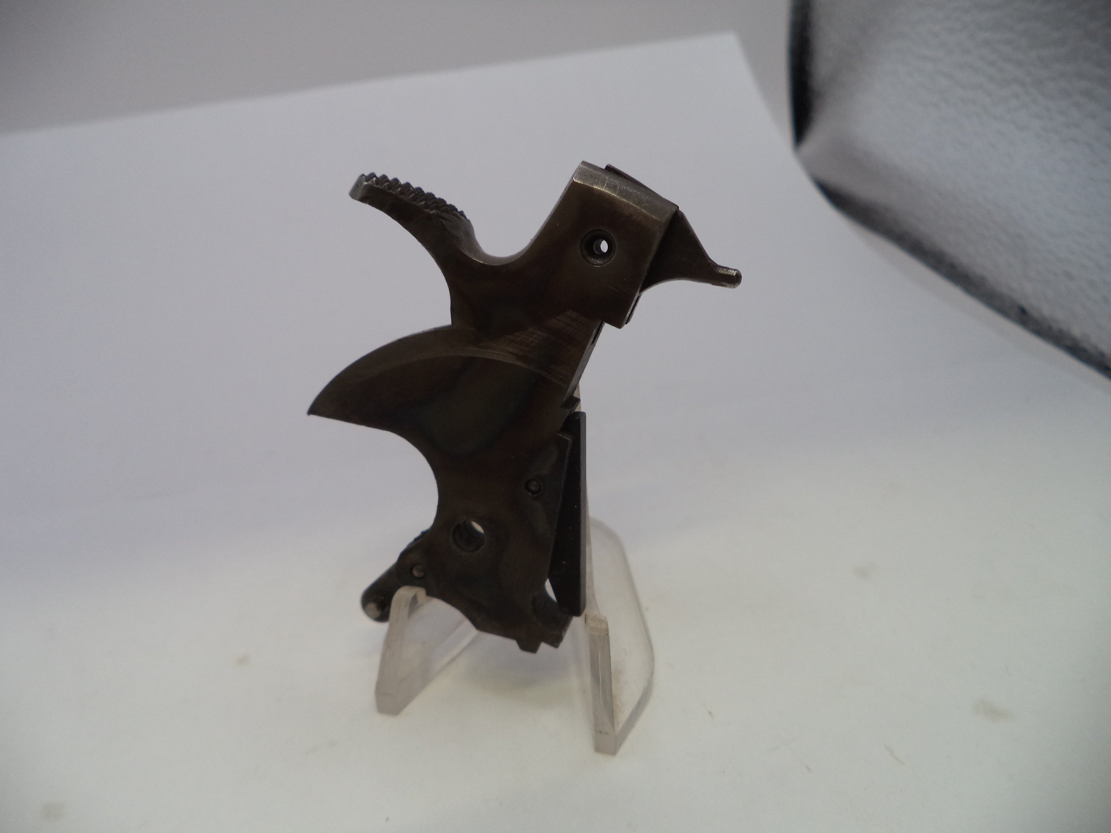 A dark brown metallic firearm magazine receiver or trigger assembly mounted on a clear plastic display stand, photographed on a white surface.