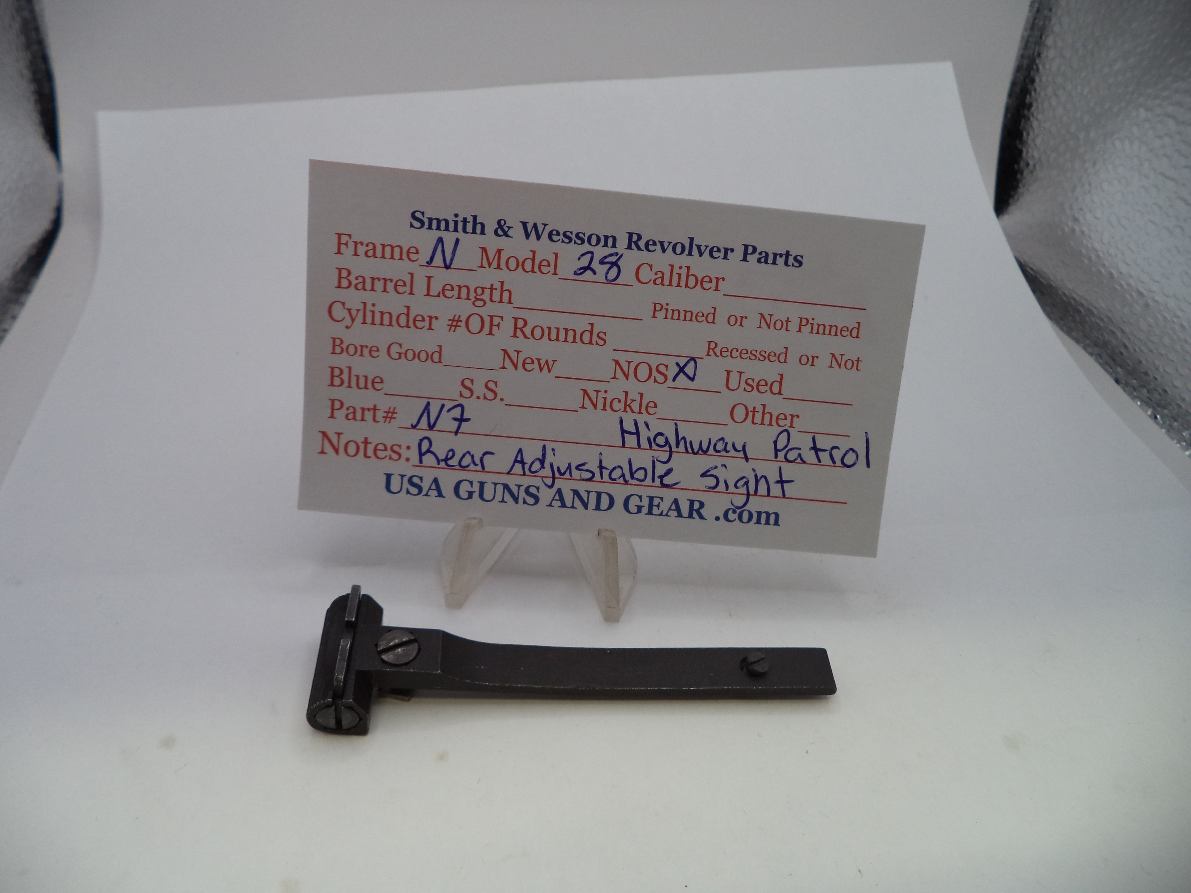A small black metal revolver part displayed on a clear stand in front of a printed parts information card for a Smith & Wesson revolver, photographed on a white surface.