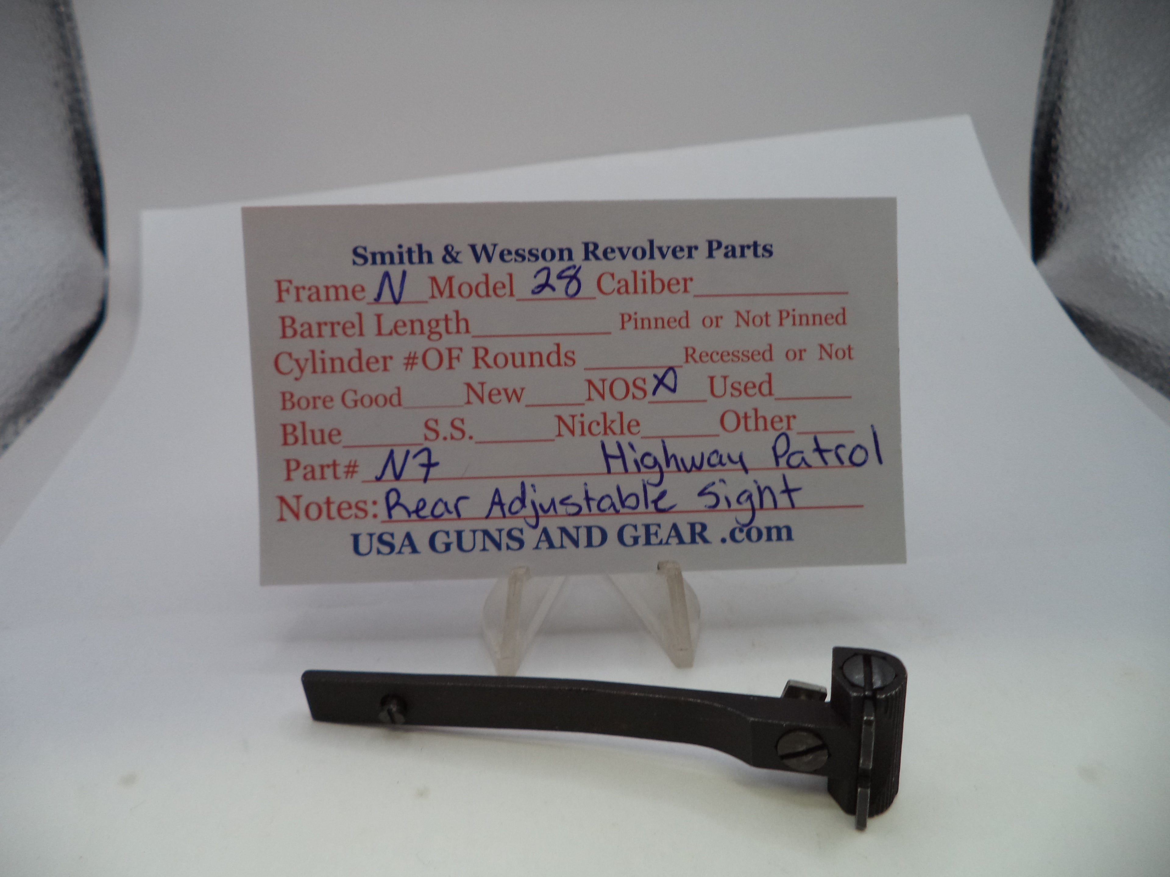 A black metal Smith & Wesson revolver part displayed on a clear stand in front of a printed parts information card on a white surface.