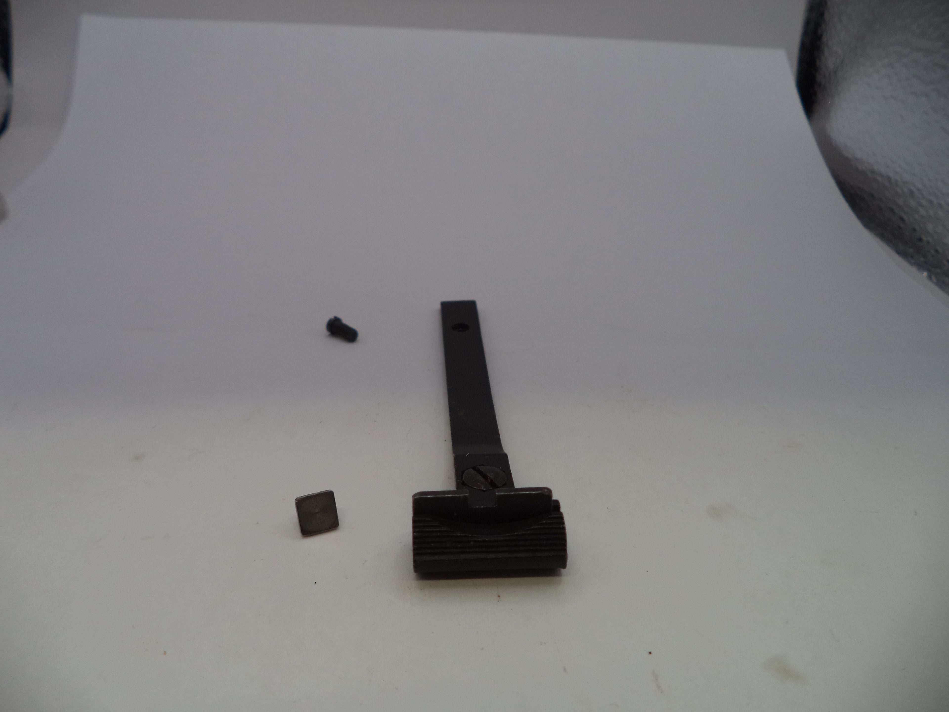 A black metal tool or hardware piece with a long handle and a textured head, shown on a white surface with two small detached parts nearby.