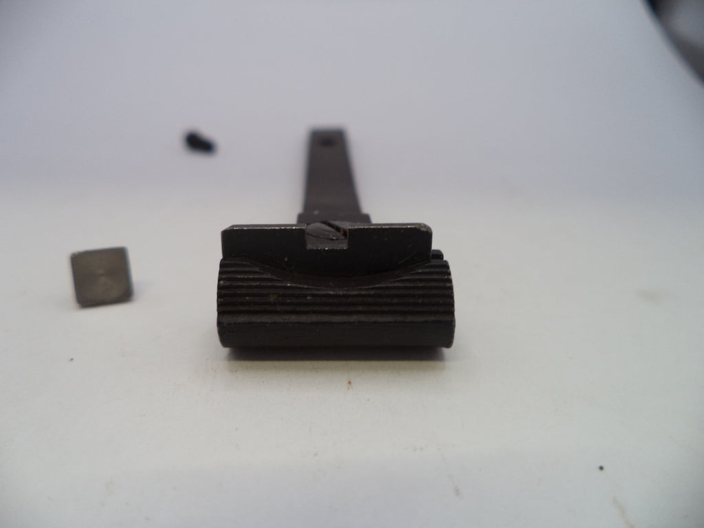 A close-up of a dark metal mechanical component or tool part on a plain light background, with a small separate square piece and a tiny dark object blurred in the background.