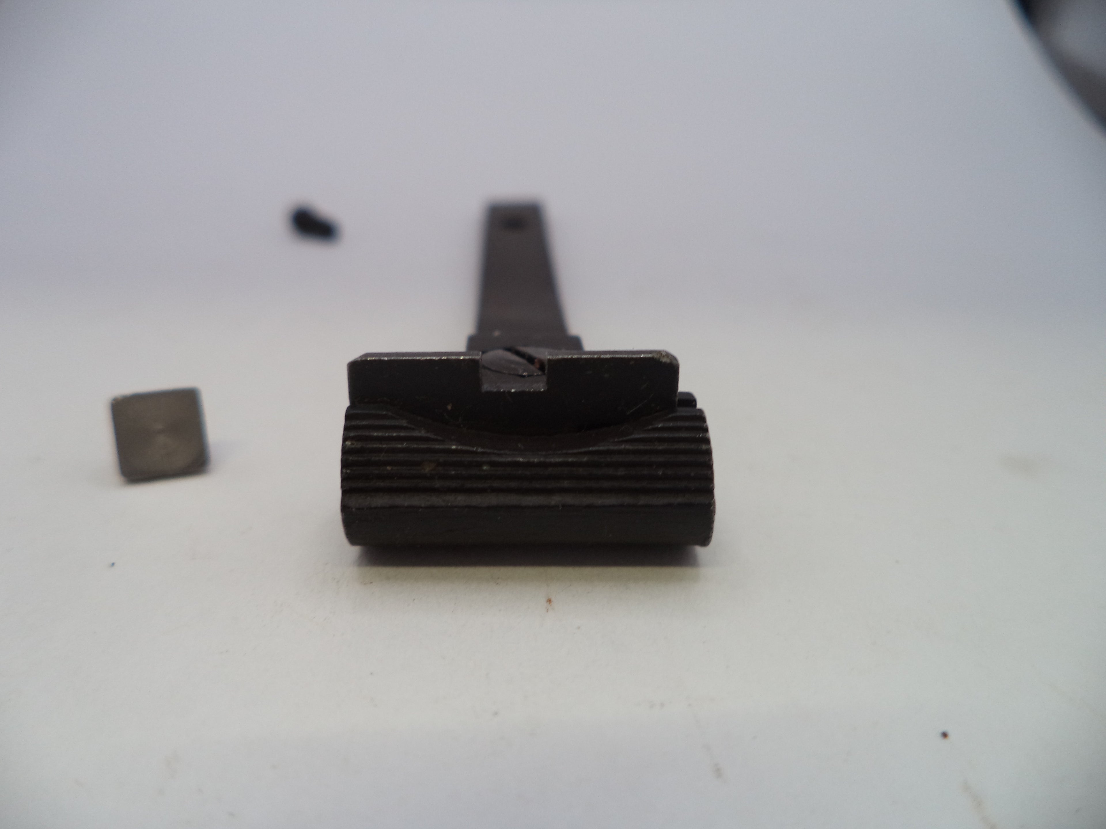 A close-up of a dark metal mechanical component or tool part on a plain light background, with a small separate square piece and a tiny dark object blurred in the background.