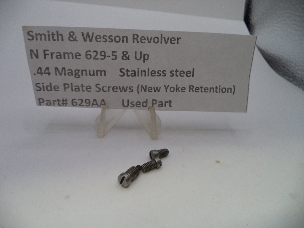Two small stainless steel side plate screws for a Smith & Wesson revolver, displayed on a white surface with a paper label card behind them.