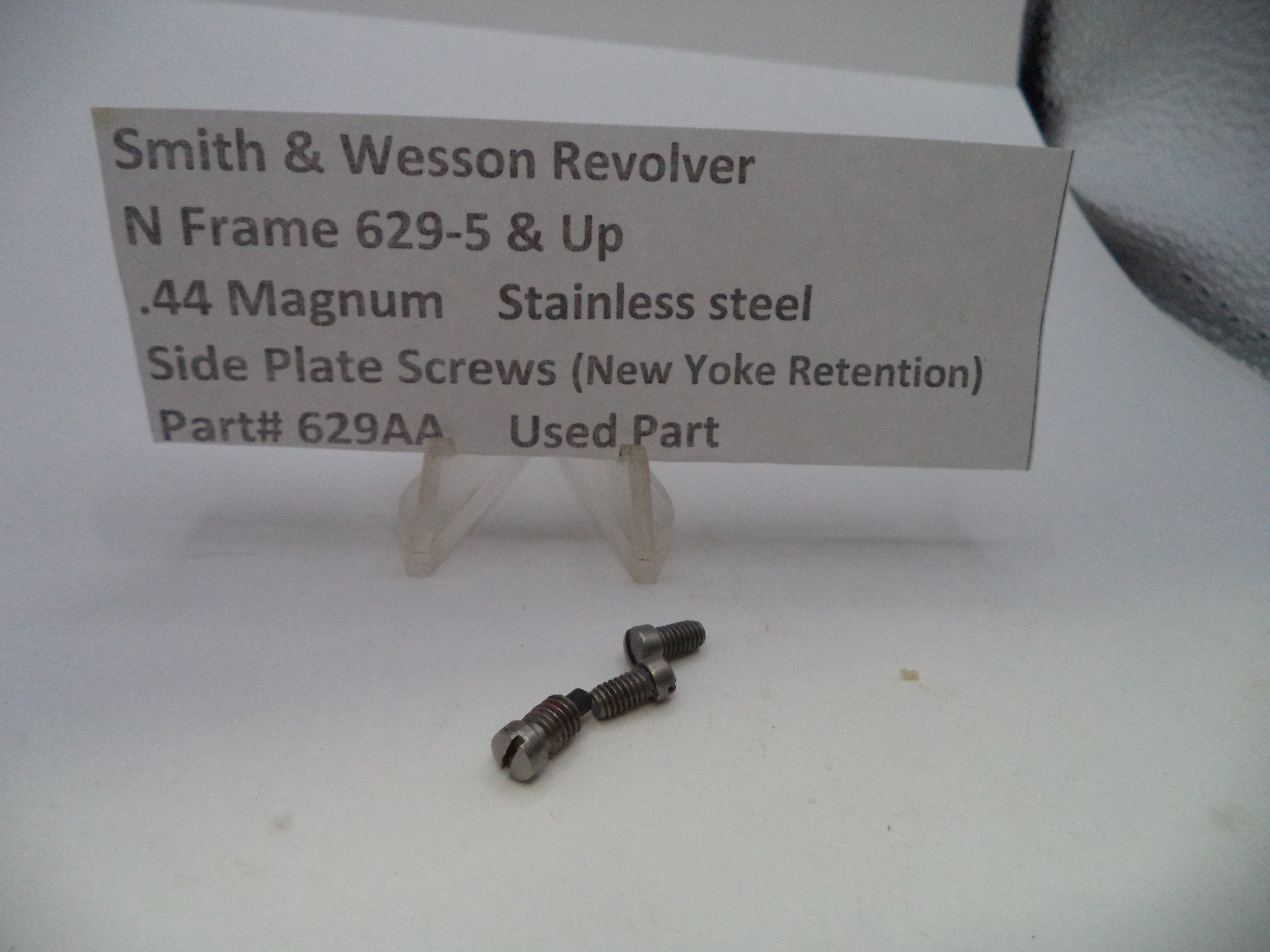Two small stainless steel side plate screws for a Smith & Wesson revolver, displayed on a white surface with a paper label card behind them.