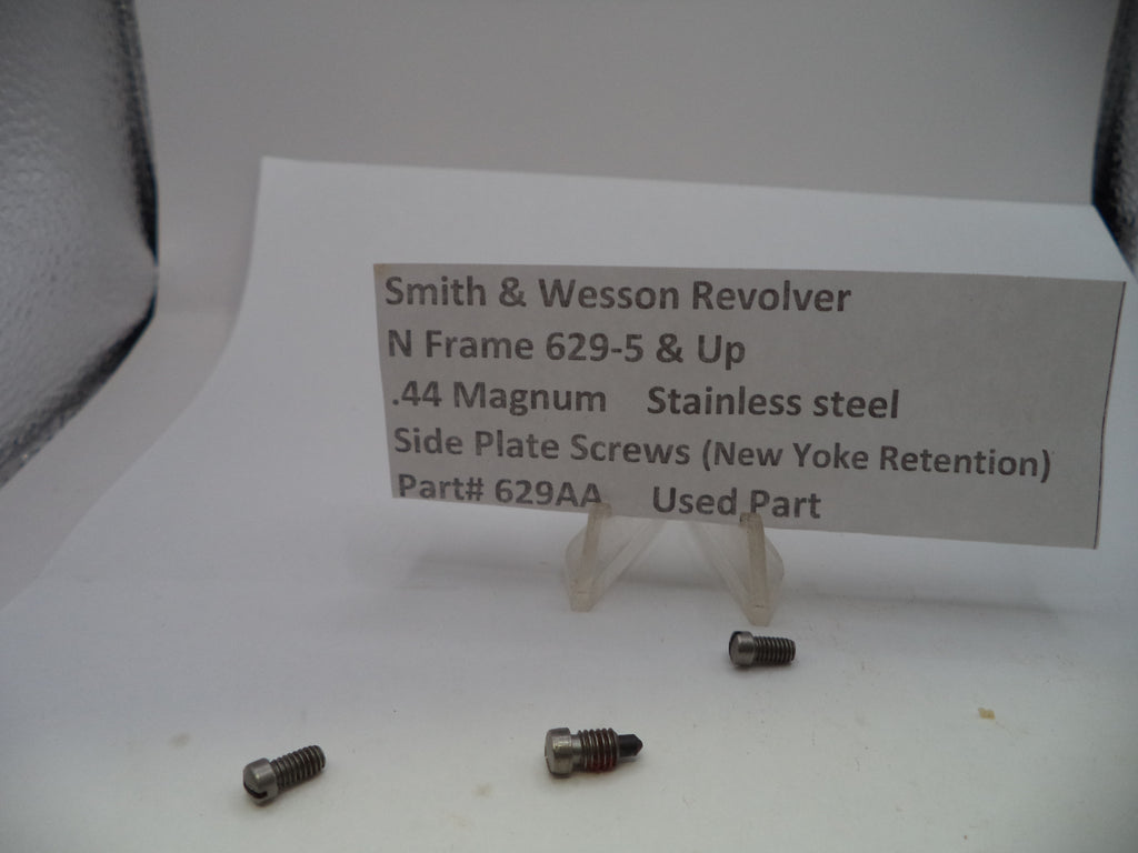 Four small stainless steel screws for a Smith & Wesson revolver, displayed on a white surface with a paper label card in the background.