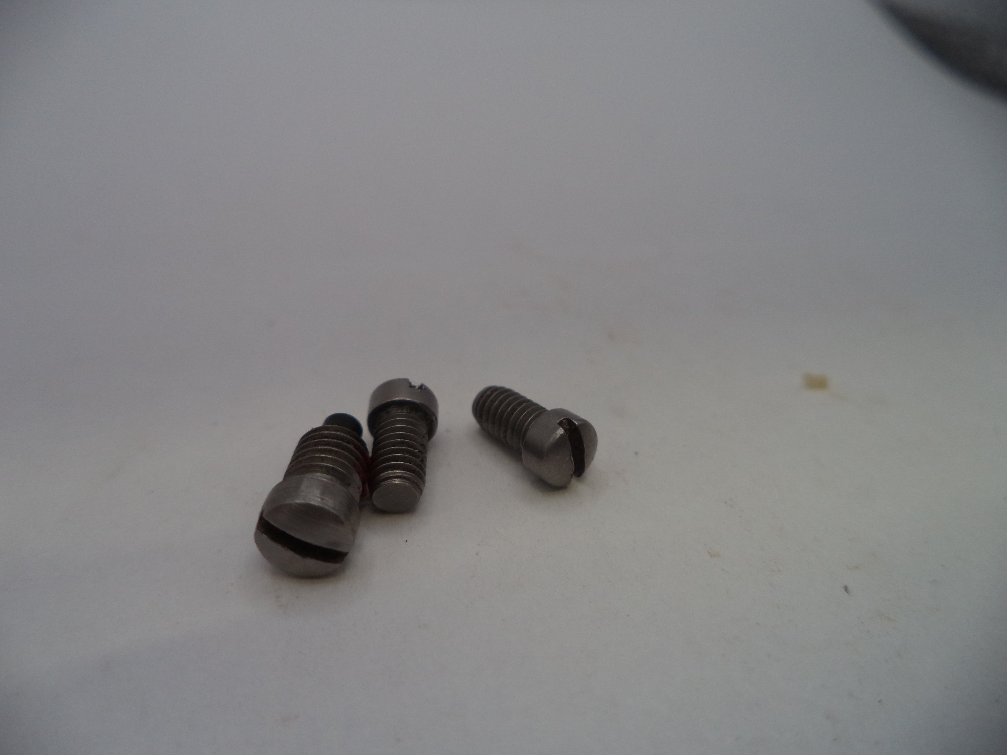 Three small metallic screws with slotted cross-drilled heads, showing a dark metallic gray finish, resting on a plain off-white surface.