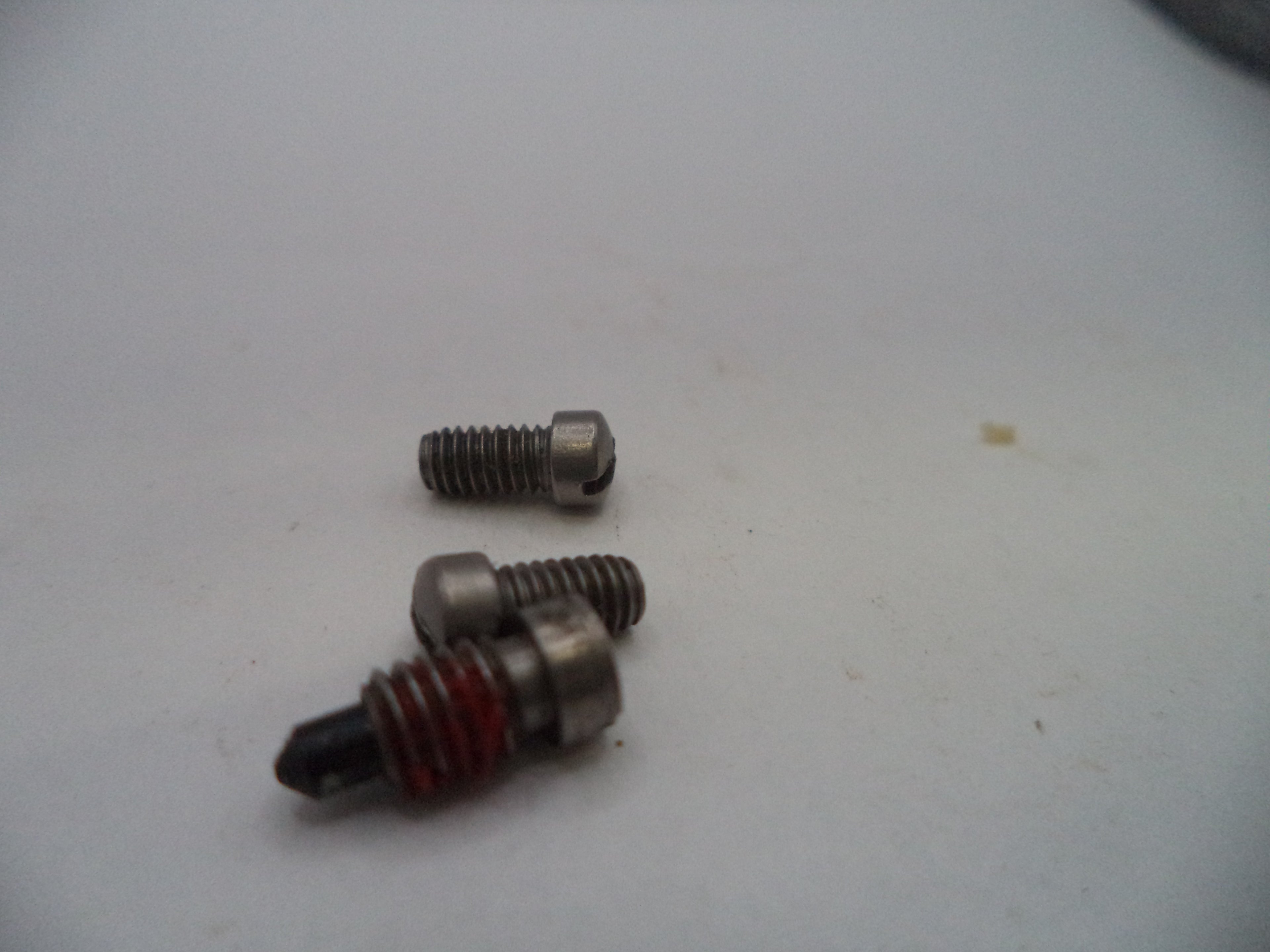 Three small metal threaded components with one featuring a red spring, photographed on a plain off-white surface.