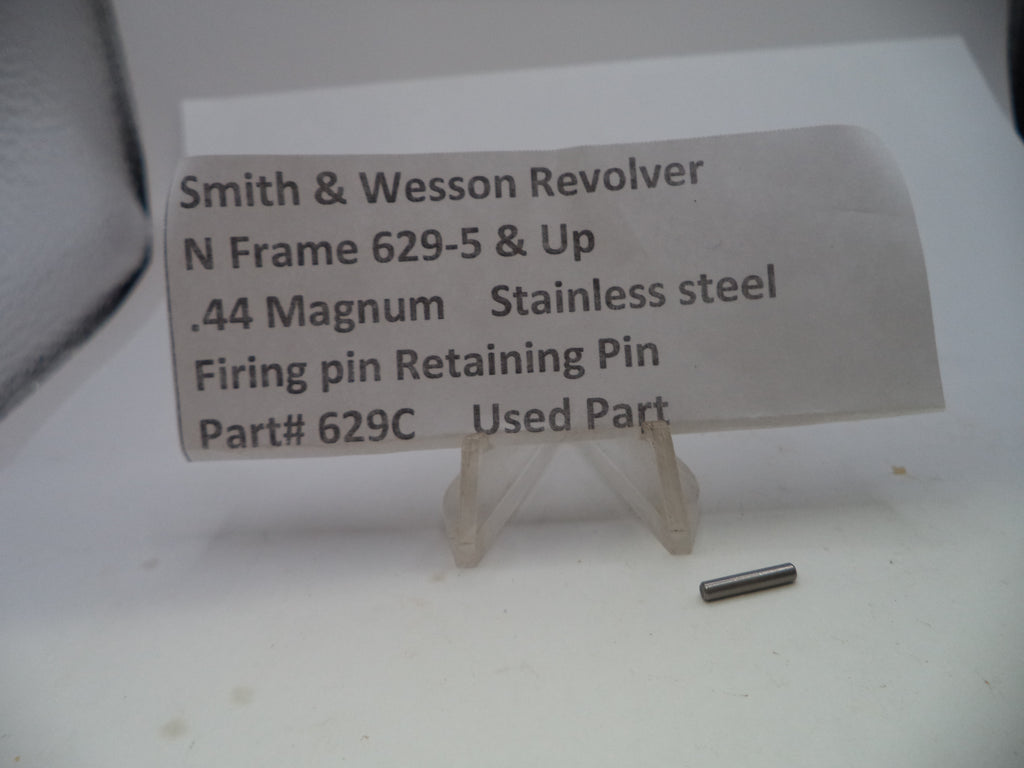 A small stainless steel firing pin retaining pin displayed on a clear plastic stand next to a paper label that reads 'Smith & Wesson Revolver N Frame 629-5 & Up .44 Magnum Stainless steel Firing pin Retaining Pin Part# 629C Used Part'.