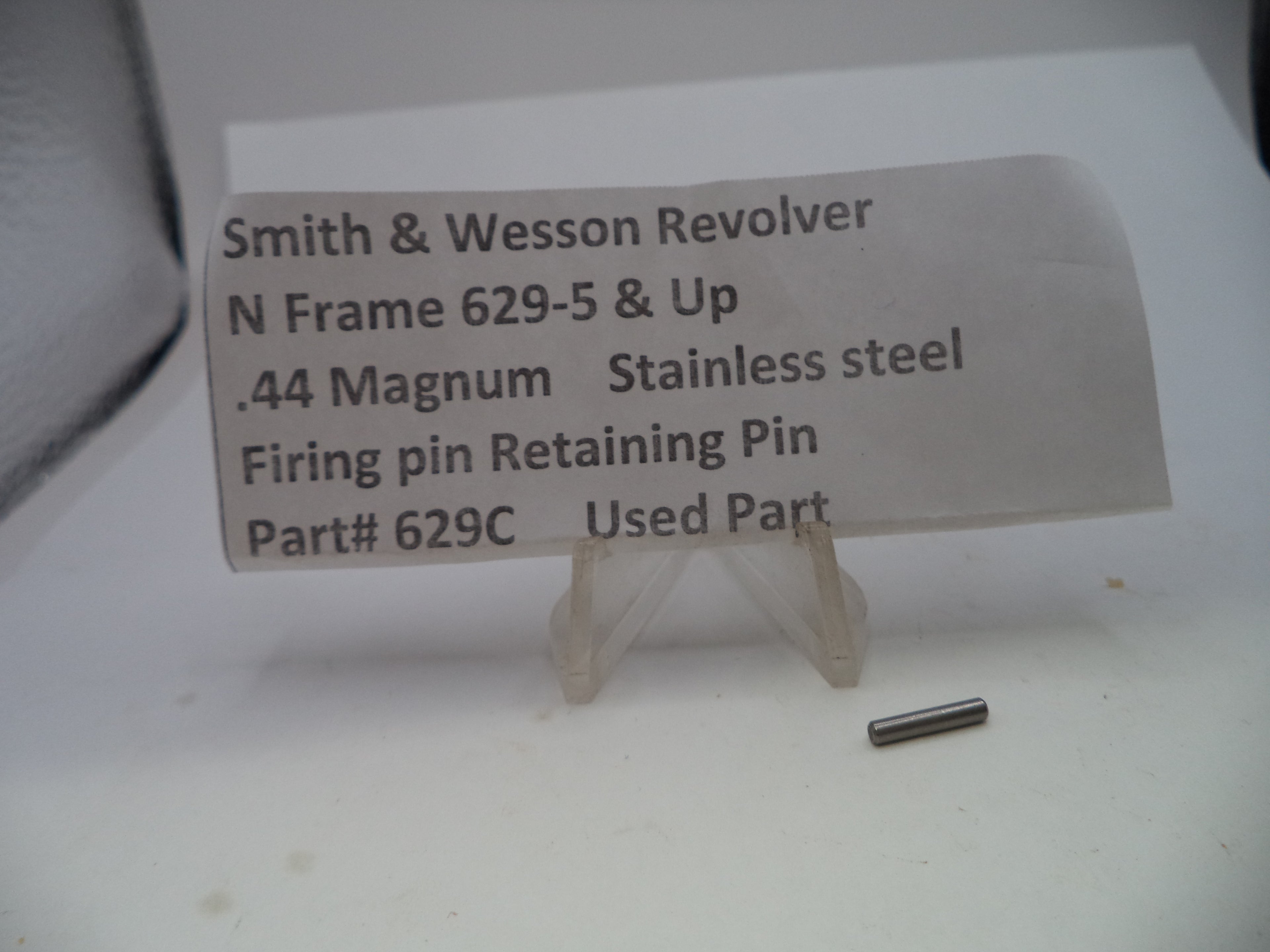 A small stainless steel firing pin retaining pin displayed on a clear plastic stand next to a paper label that reads 'Smith & Wesson Revolver N Frame 629-5 & Up .44 Magnum Stainless steel Firing pin Retaining Pin Part# 629C Used Part'.