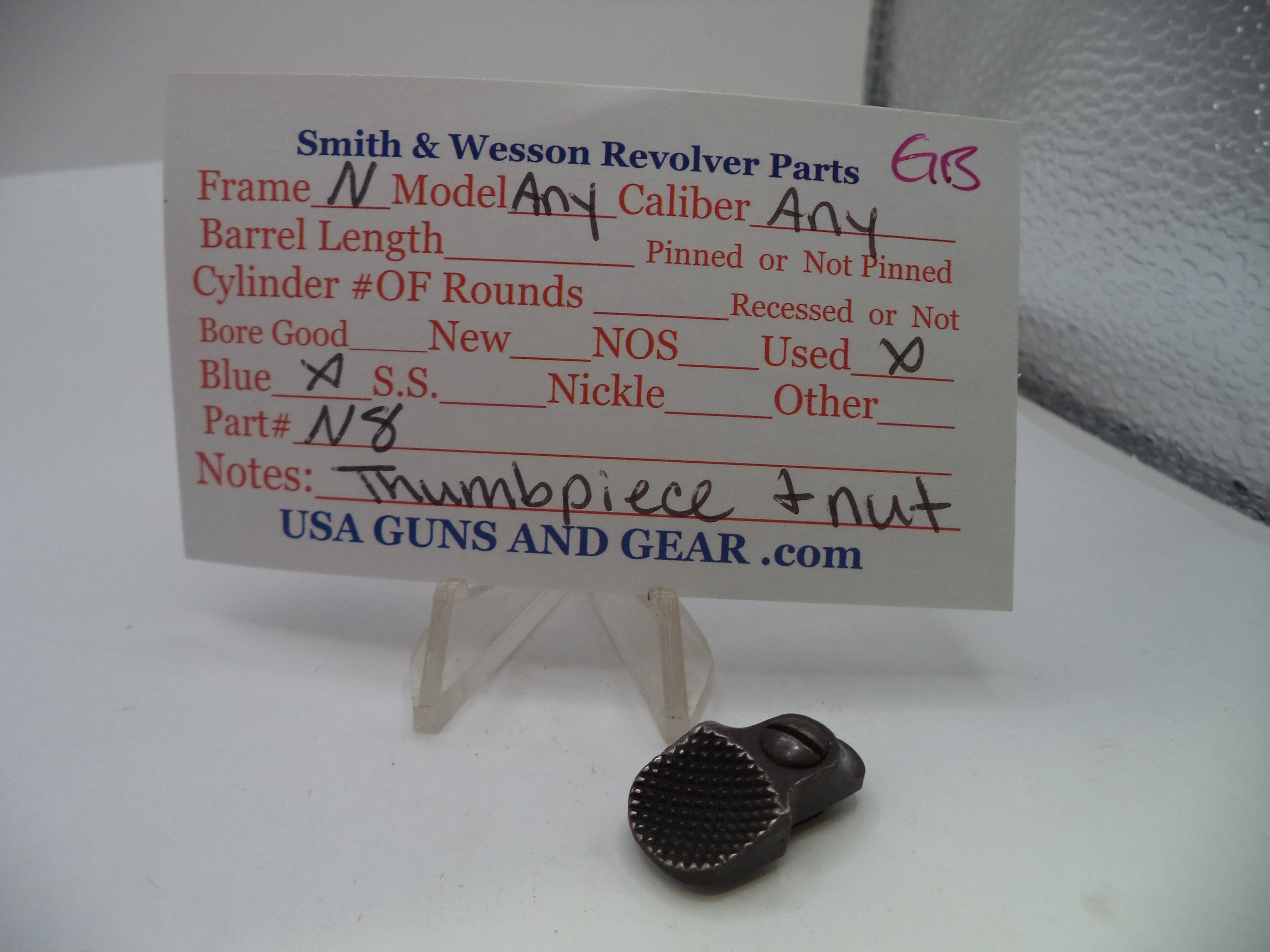 A small dark metal revolver part displayed in front of a handwritten parts list card on a clear stand. The card reads Smith & Wesson Revolver Parts and includes fields for caliber, barrel length, and part details, with USA GUNS AND GEAR .